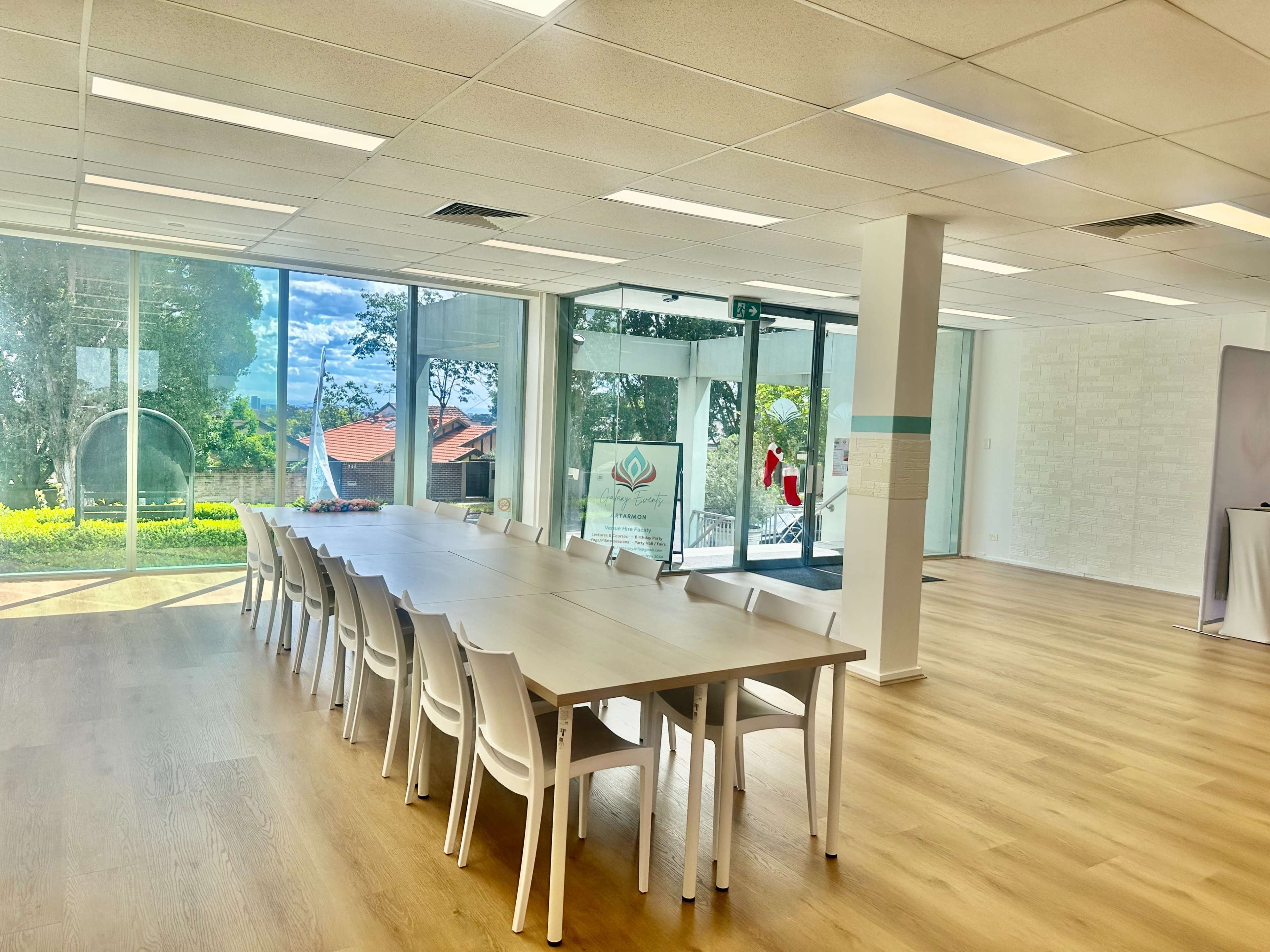 The image shows a spacious, well-lit meeting room with a long table surrounded by white chairs and large windows overlooking greenery.