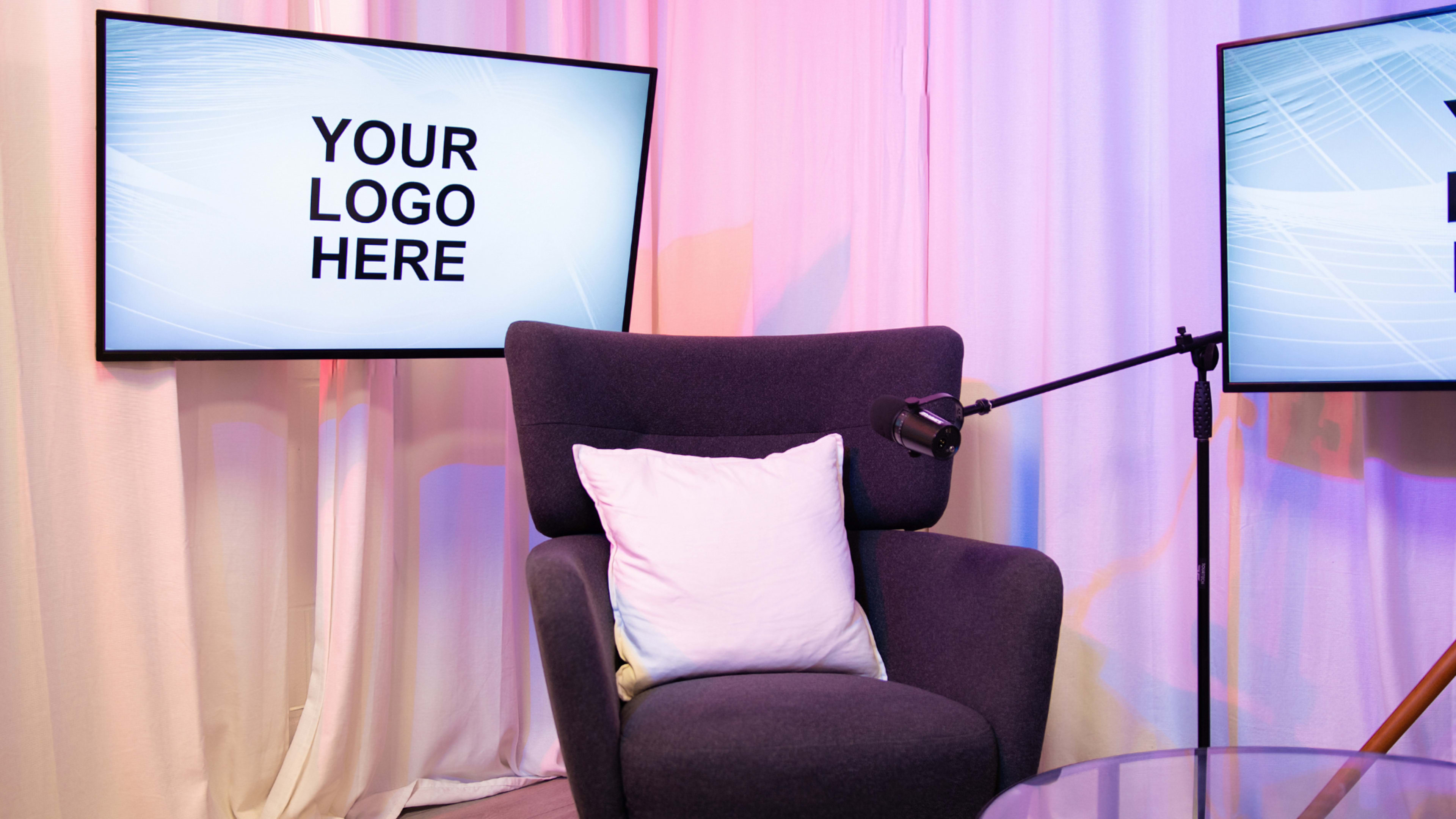 A cozy chair is positioned in front of two screens displaying the text "YOUR LOGO HERE" within a softly lit, professionally designed space.