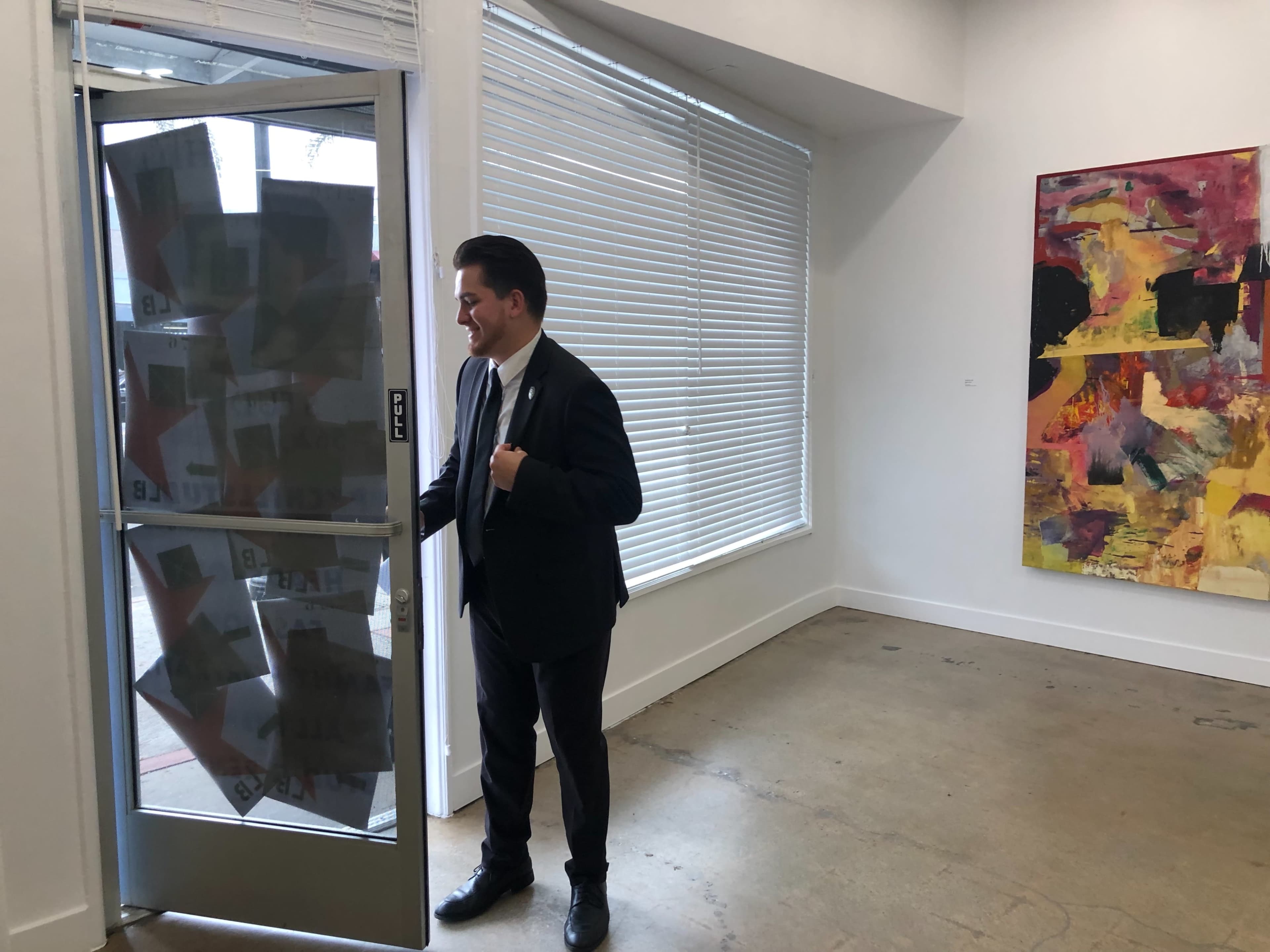 A man in a suit stands by an open glass door, with signs taped to the door, in a gallery-like interior featuring a large abstract painting on the wall.