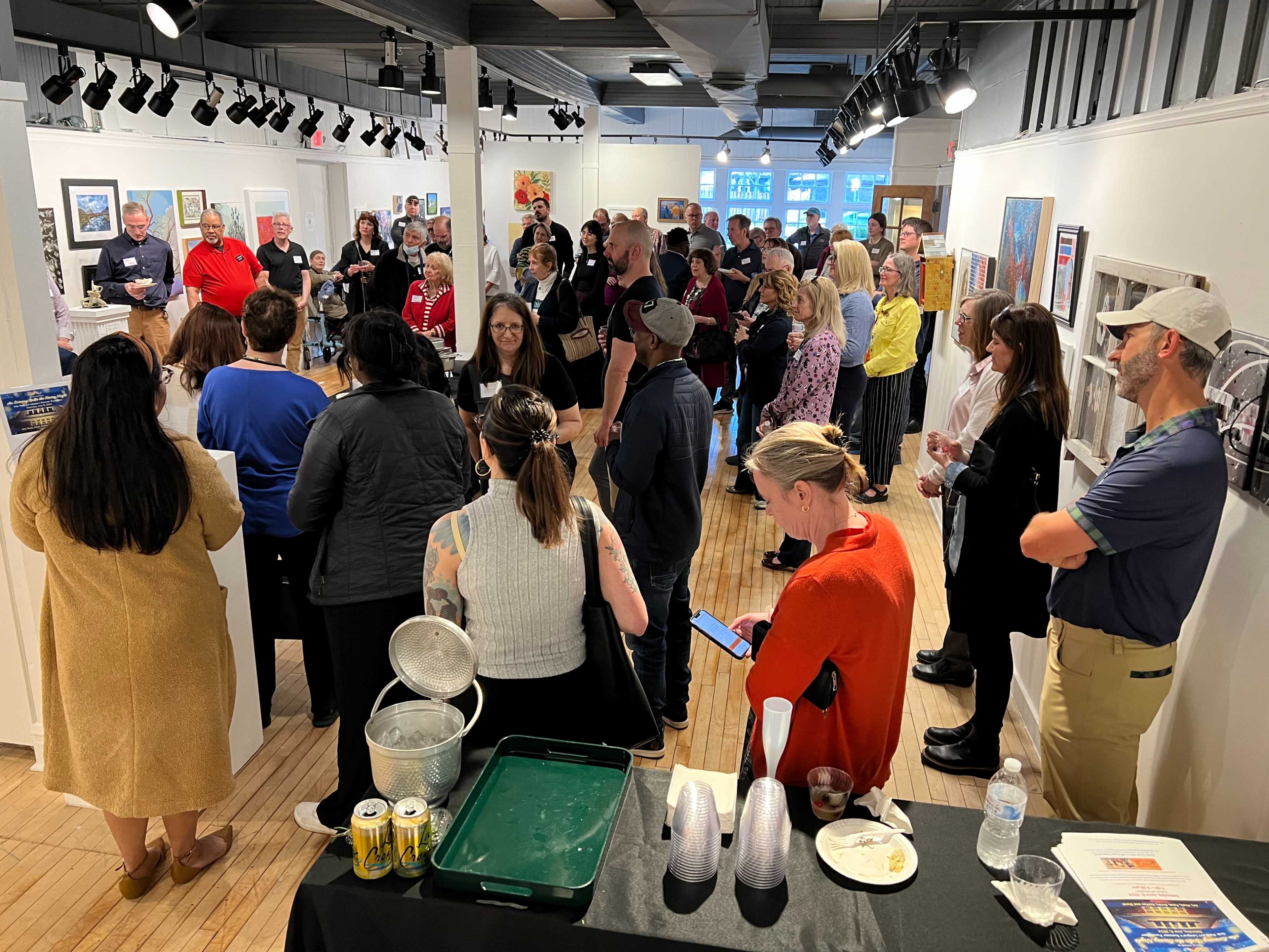 A diverse group of people gathers in an art gallery, engaging in conversation while viewing various artworks on the walls.