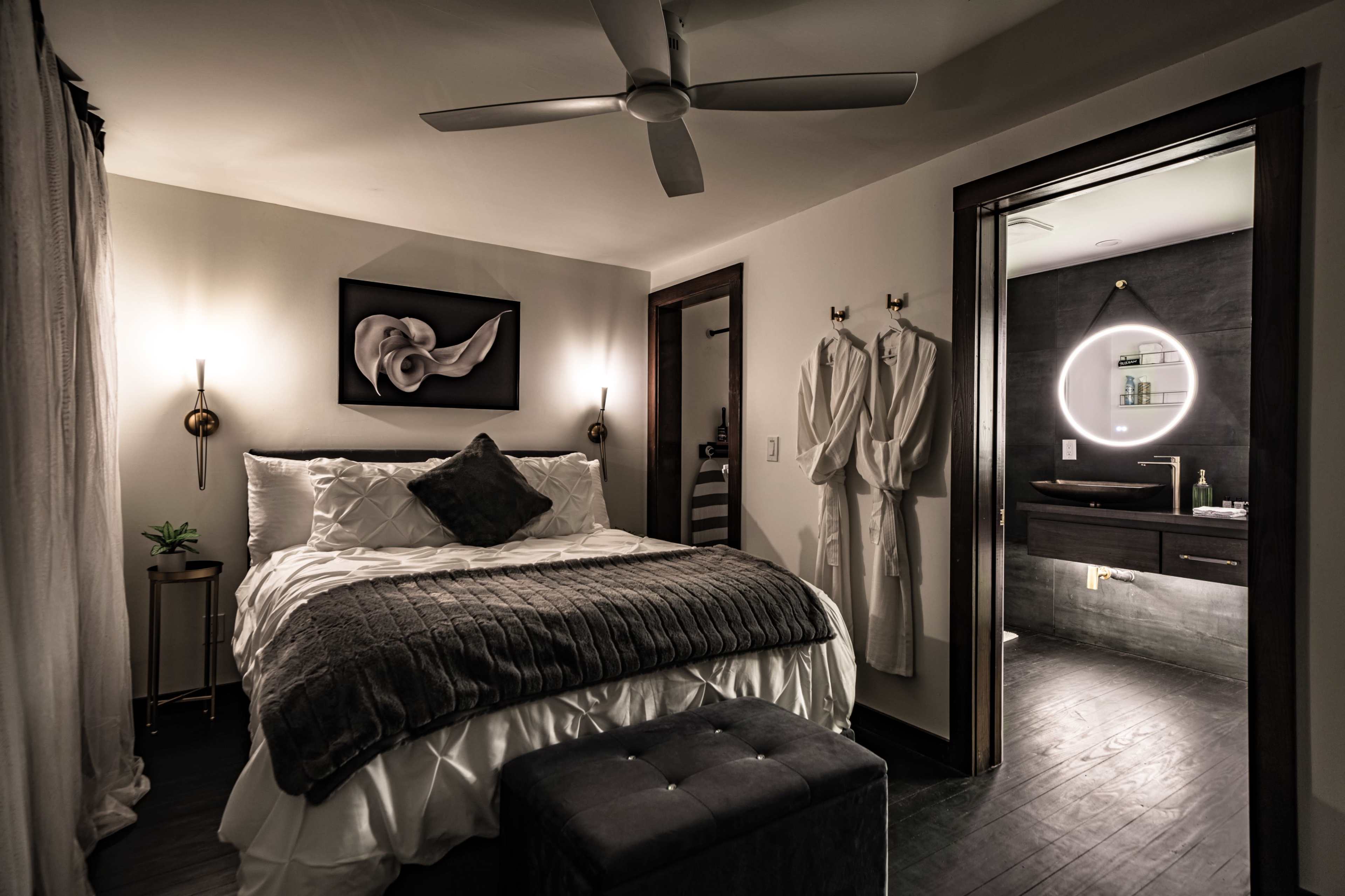 The image shows a modern bedroom with a bed covered in white linens, a nightstand with a plant, and an adjoining bathroom illuminated by a circular mirror light.