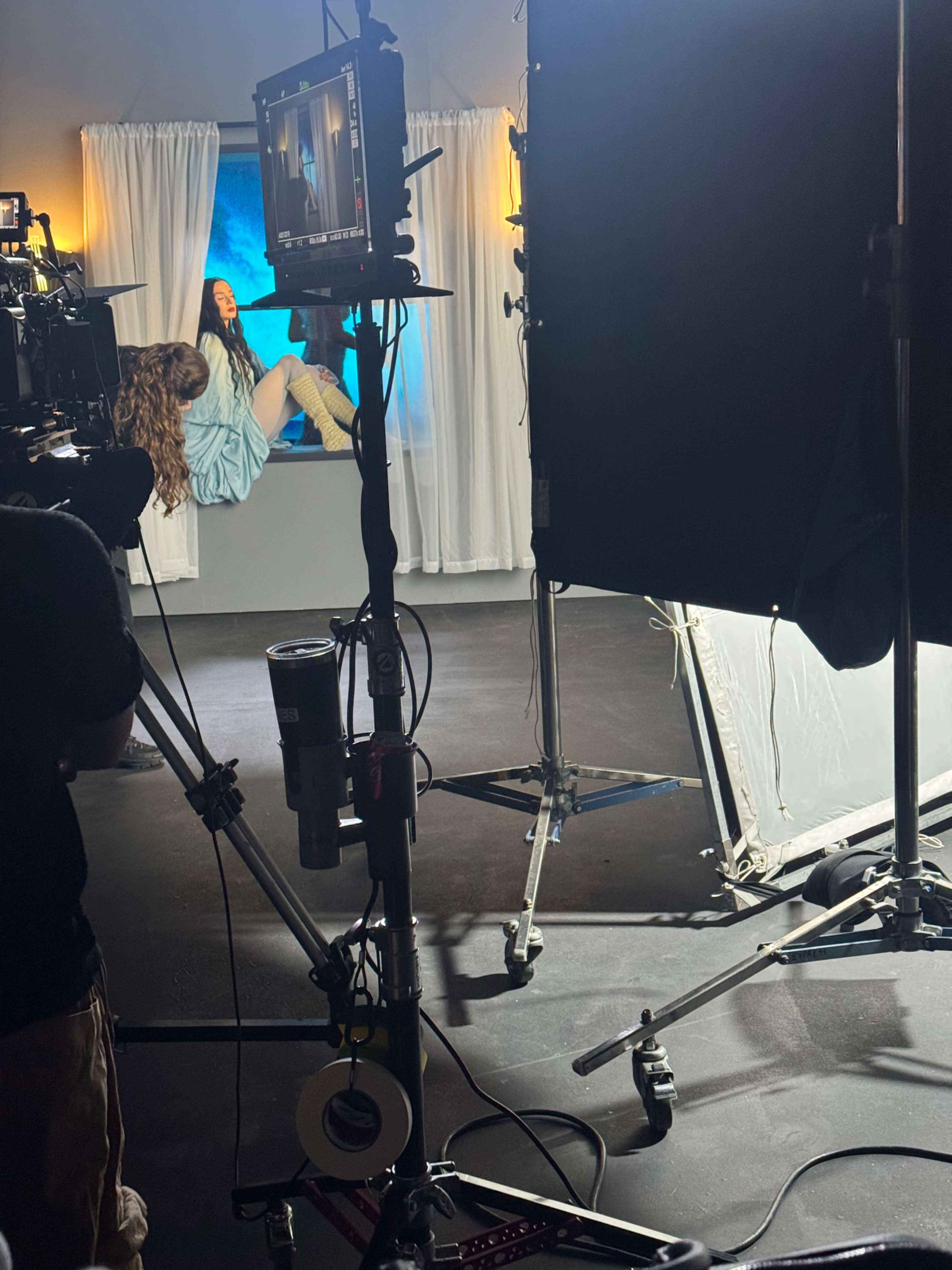 A film set is shown with a camera pointed at a model sitting on a pedestal, surrounded by lights and draped curtains.