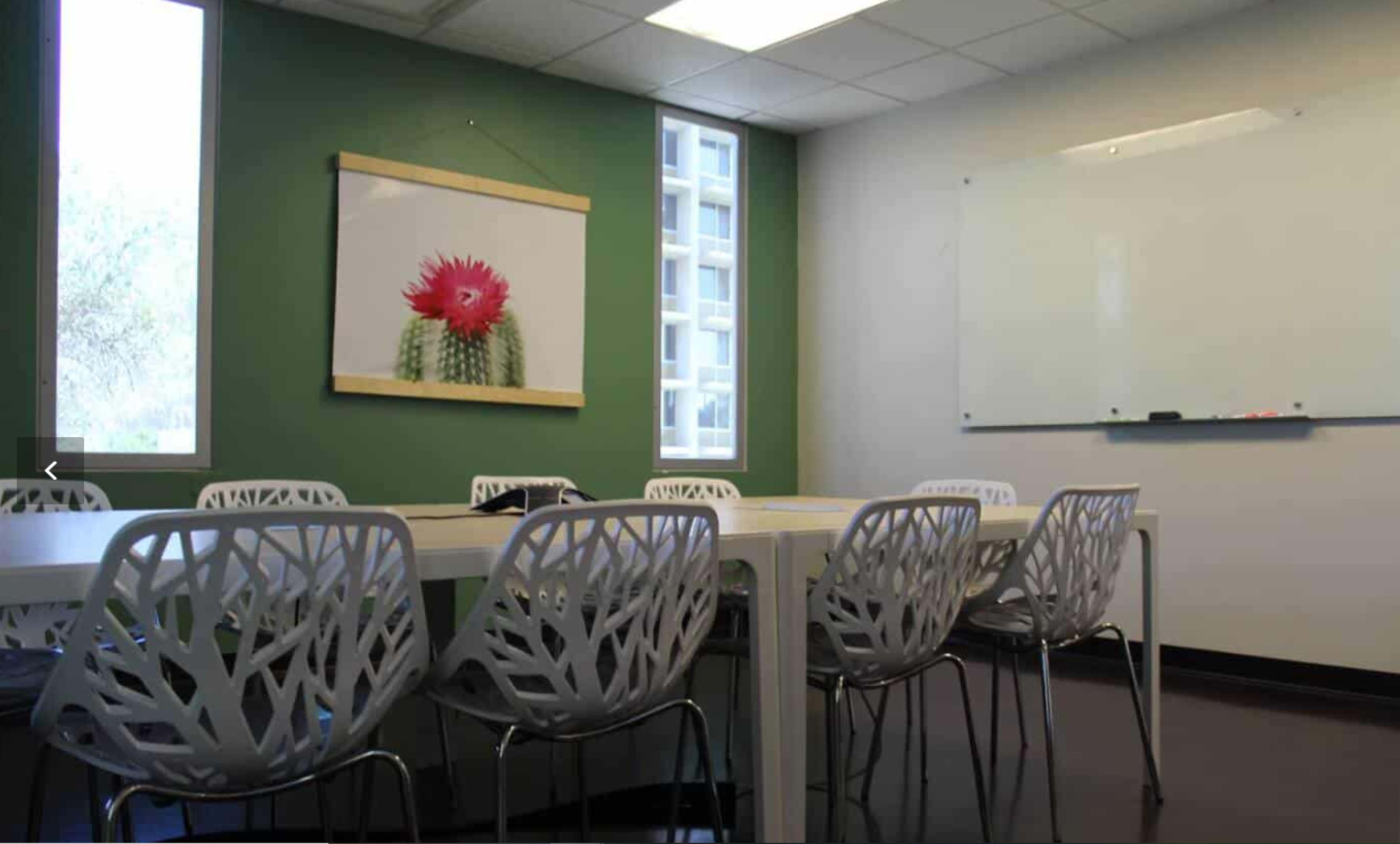 A meeting room features a white table surrounded by decorative chairs, with a large cactus print on the wall and a whiteboard nearby.