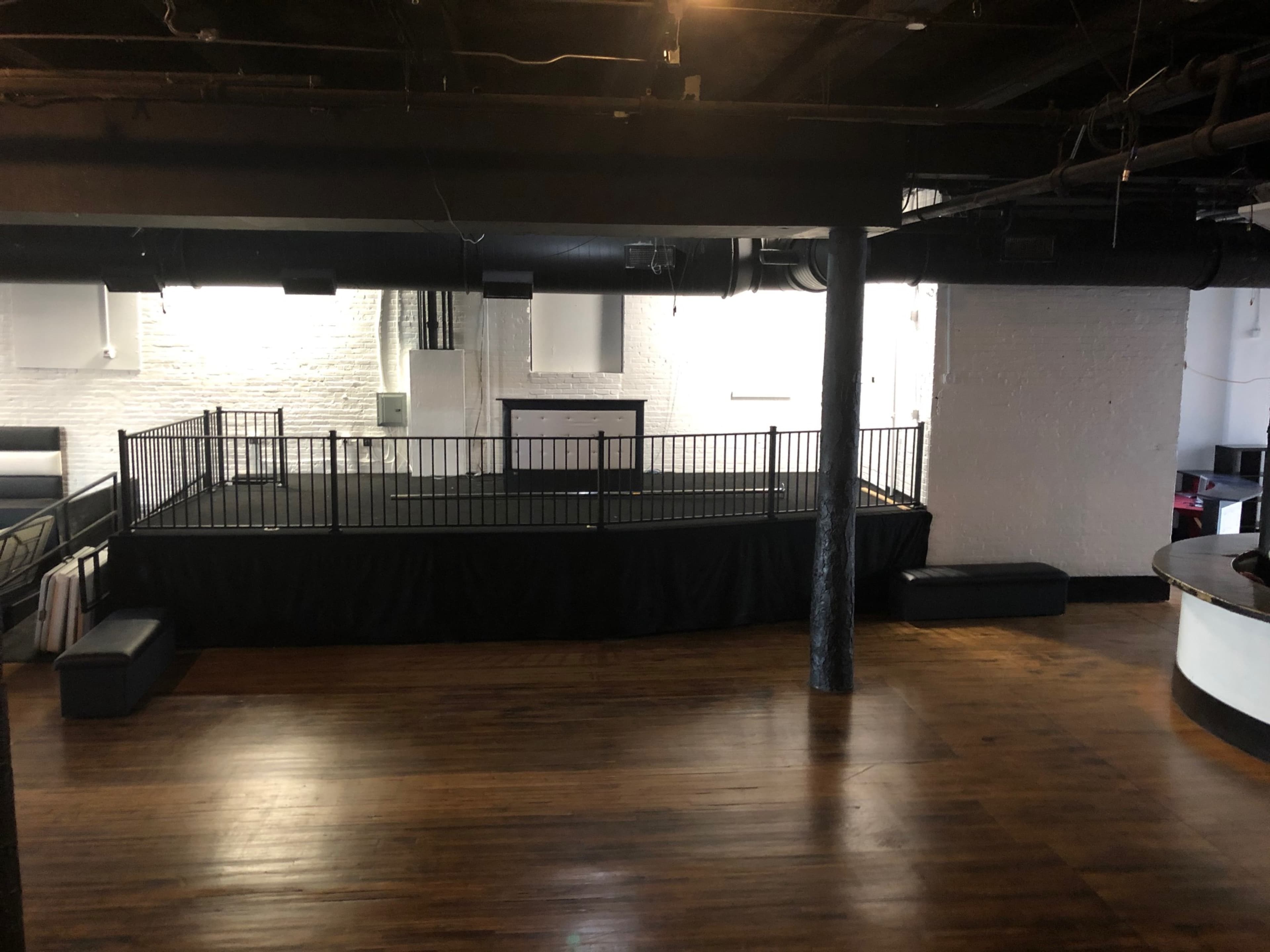 The image shows an empty, industrial-style interior space with a wooden floor, a raised black railing area, and white brick walls.