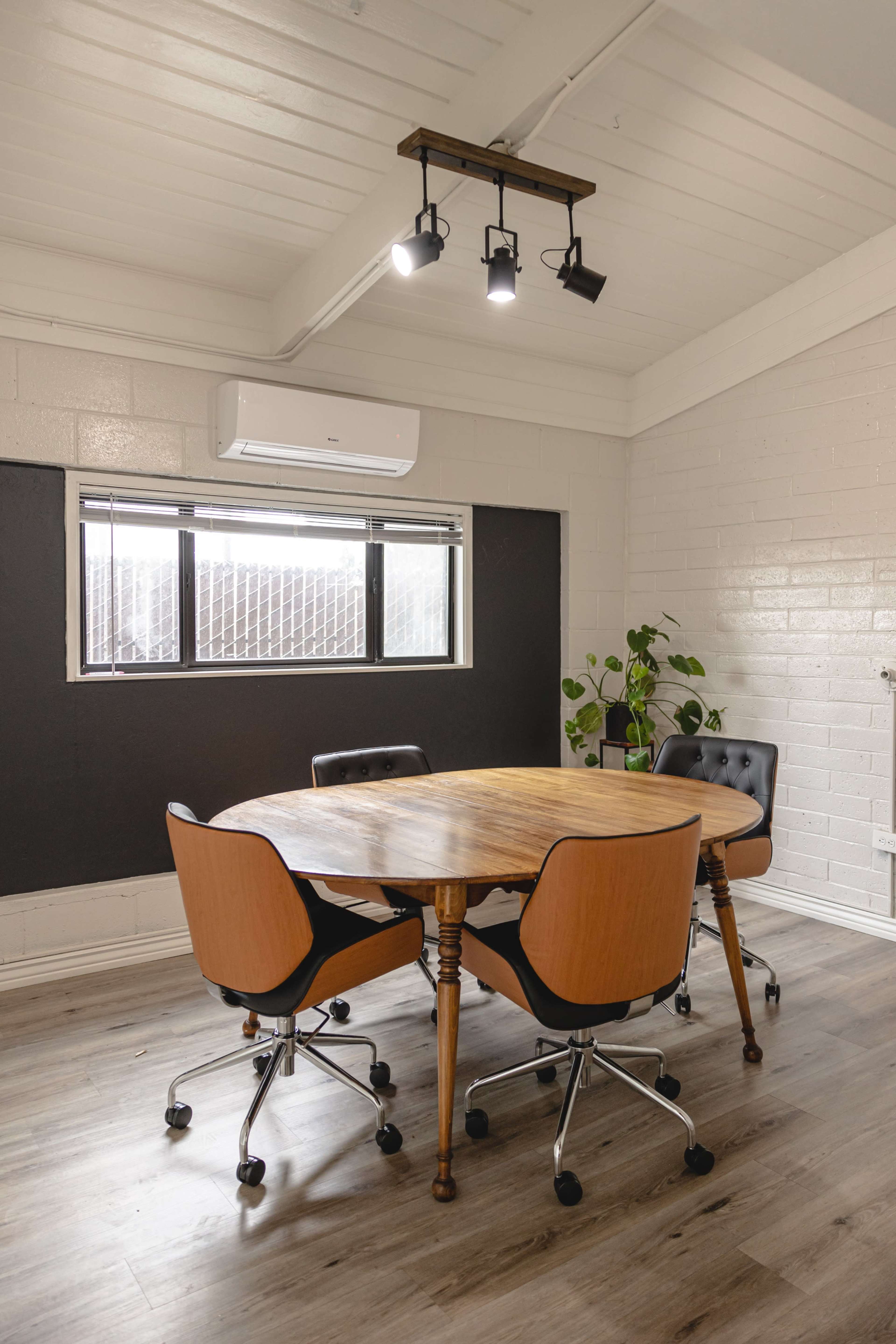 A round wooden table with four black and wooden chairs is set in a well-lit meeting room featuring a large window and a potted plant.