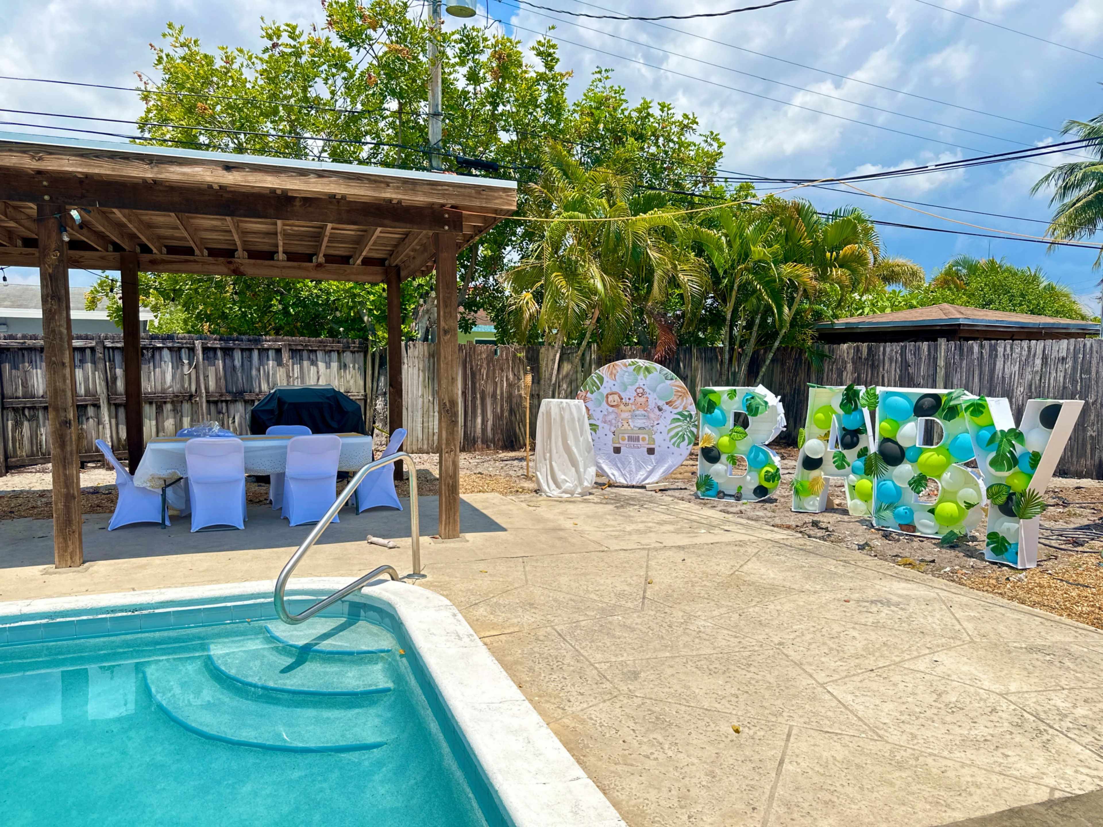 A backyard pool area with a wooden gazebo, seating arrangements under it, and a large decorative sign spelling "BABY" adorned with colorful balloons.