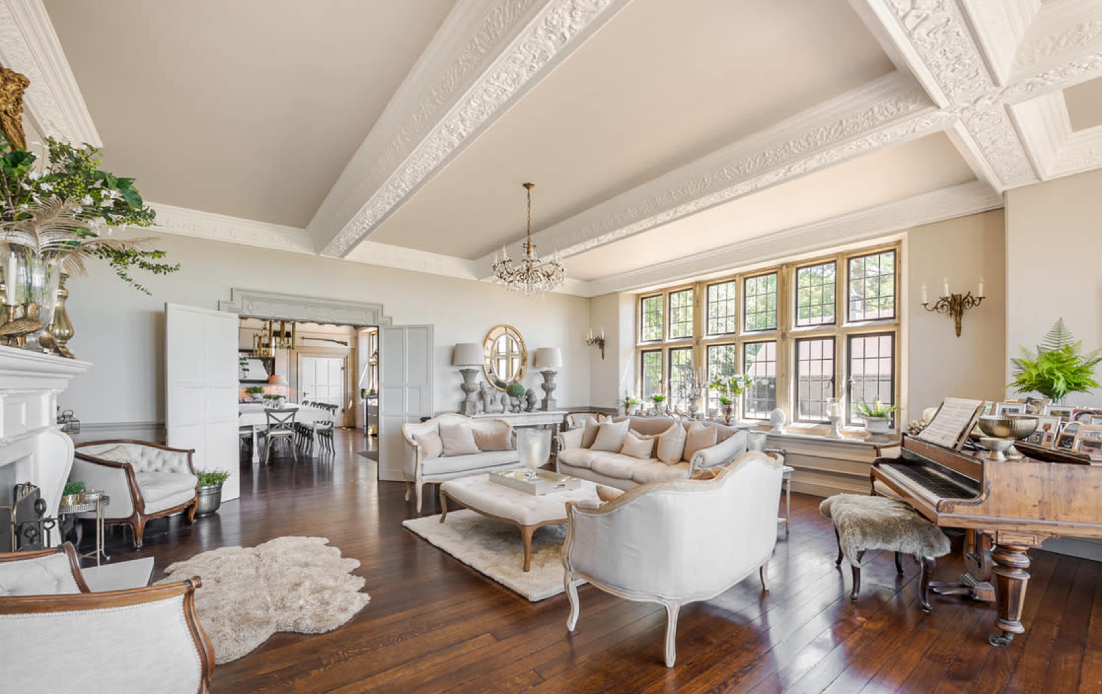 A spacious living room features elegant furniture, large windows, and a grand piano, highlighted by intricate ceiling details and warm wooden floors.
