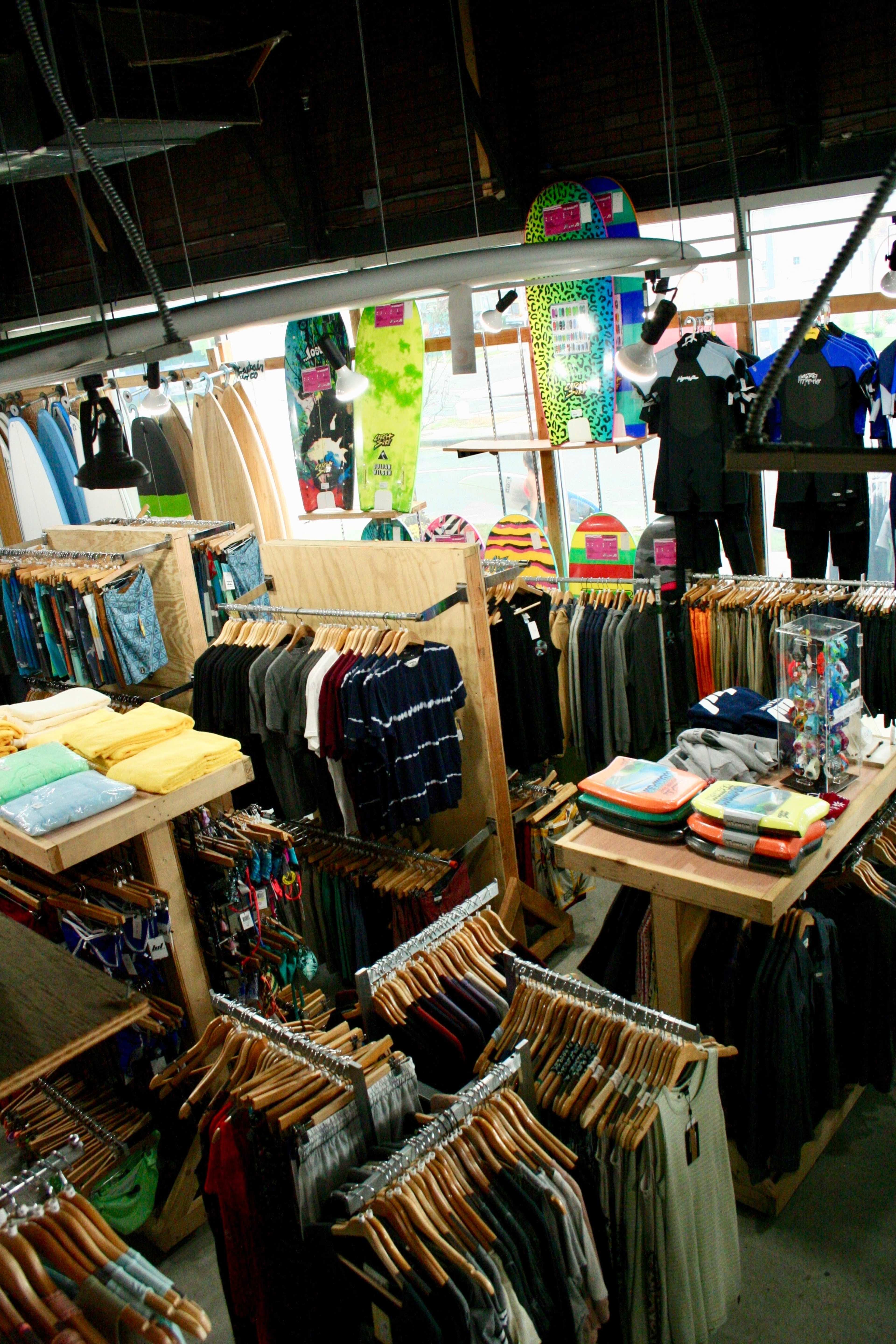 The image shows a retail store displaying clothing on wooden racks, surfboards hanging above, and various accessories arranged on tables.