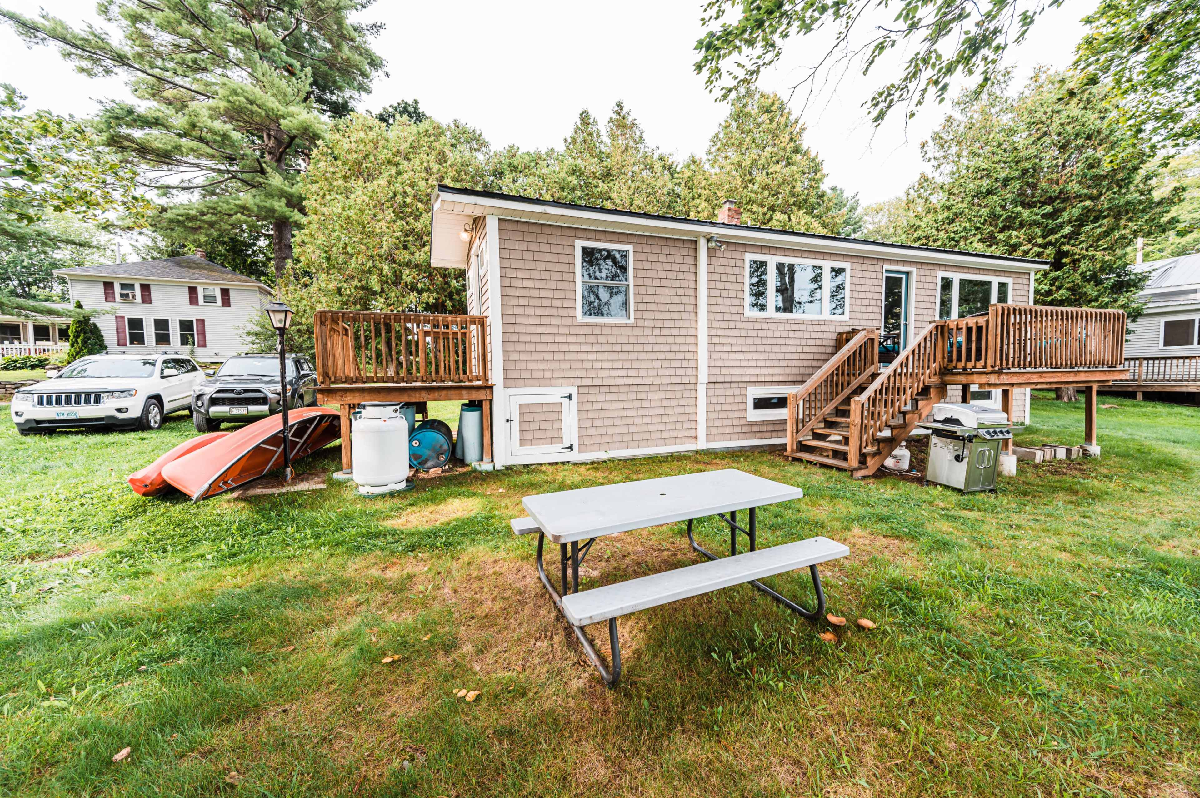 A beige house with a wooden deck and picnic table is set in a grassy backyard, flanked by trees and parked cars.