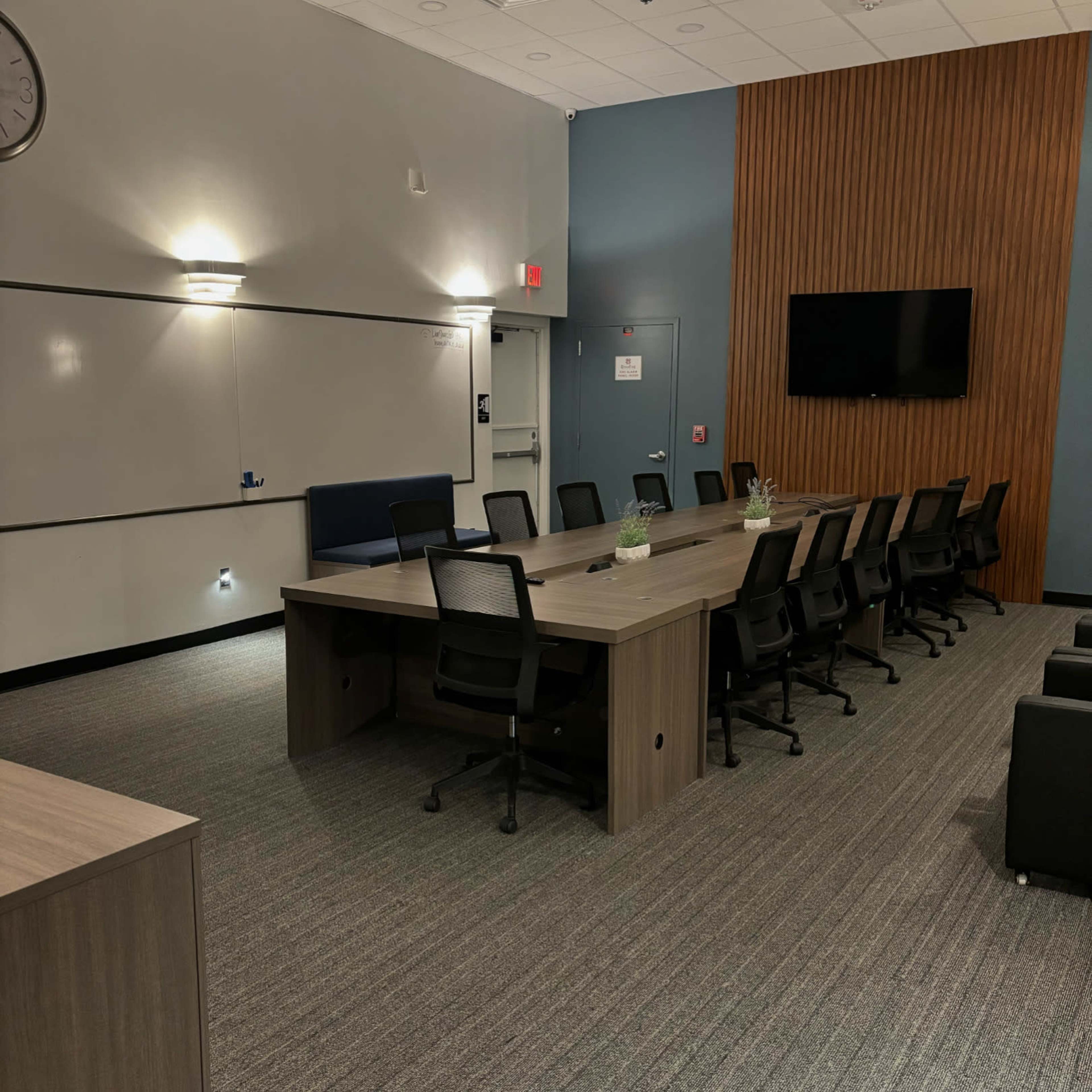 A modern conference room features a long wooden table surrounded by black chairs, with a large wall-mounted TV and a presentation board.