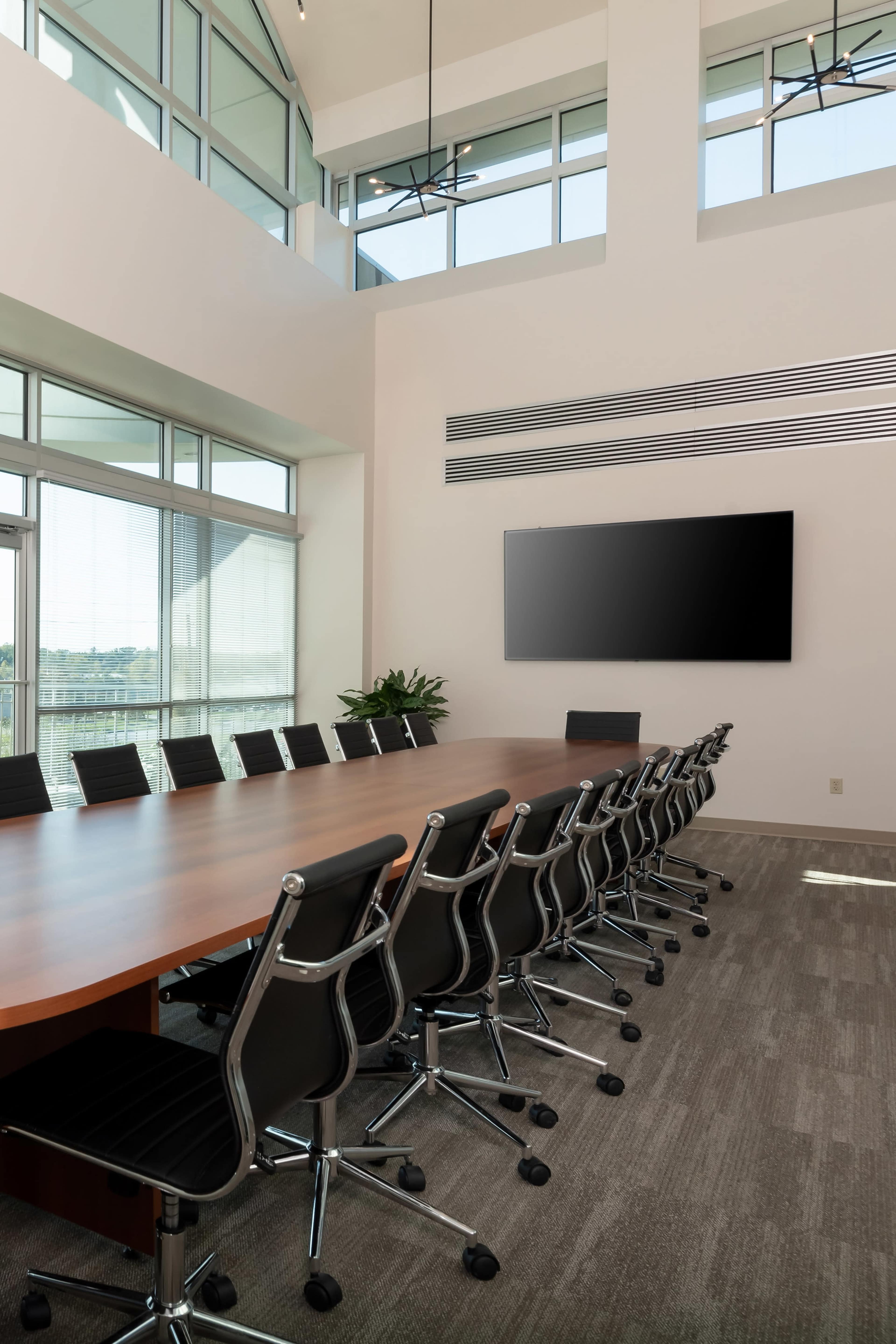 A large conference room features a long wooden table surrounded by black swivel chairs, with floor-to-ceiling windows letting in natural light and a wall-mounted television.