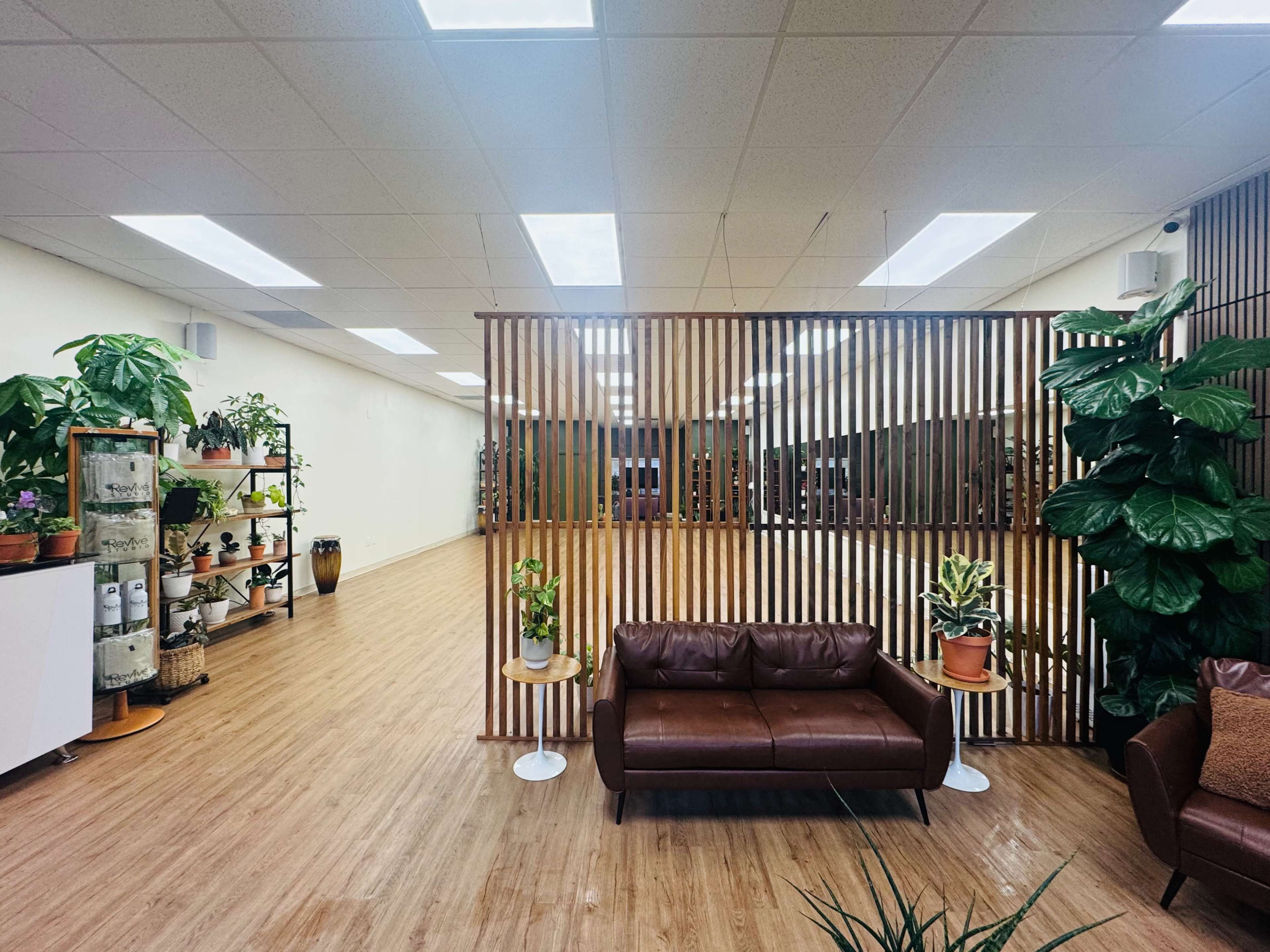 A spacious interior with wooden flooring features a brown leather sofa, a wooden divider, and potted plants along the walls.