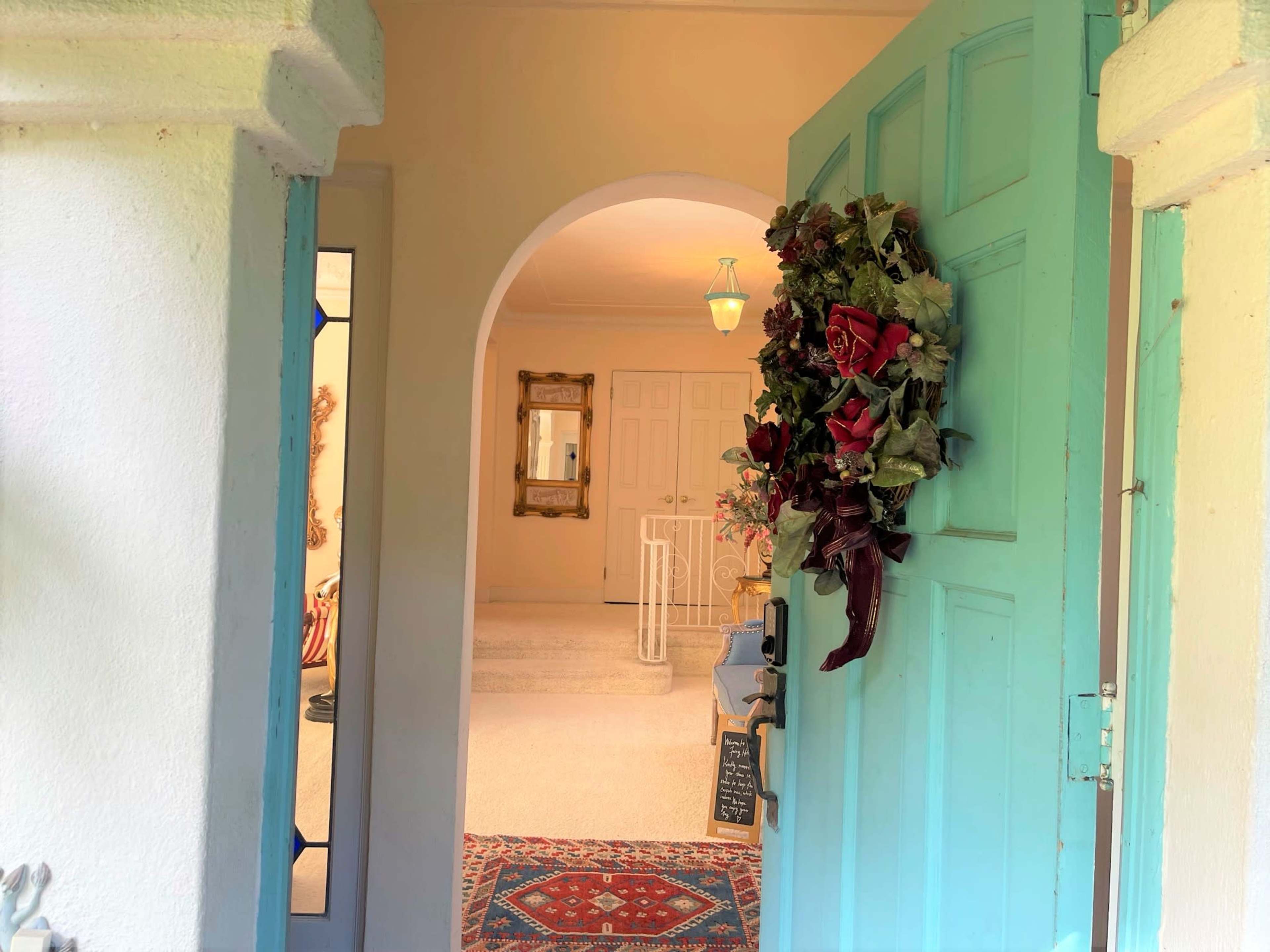 A turquoise door is open, revealing a bright hallway with a decorative mirror and a floral wreath adorning the door.