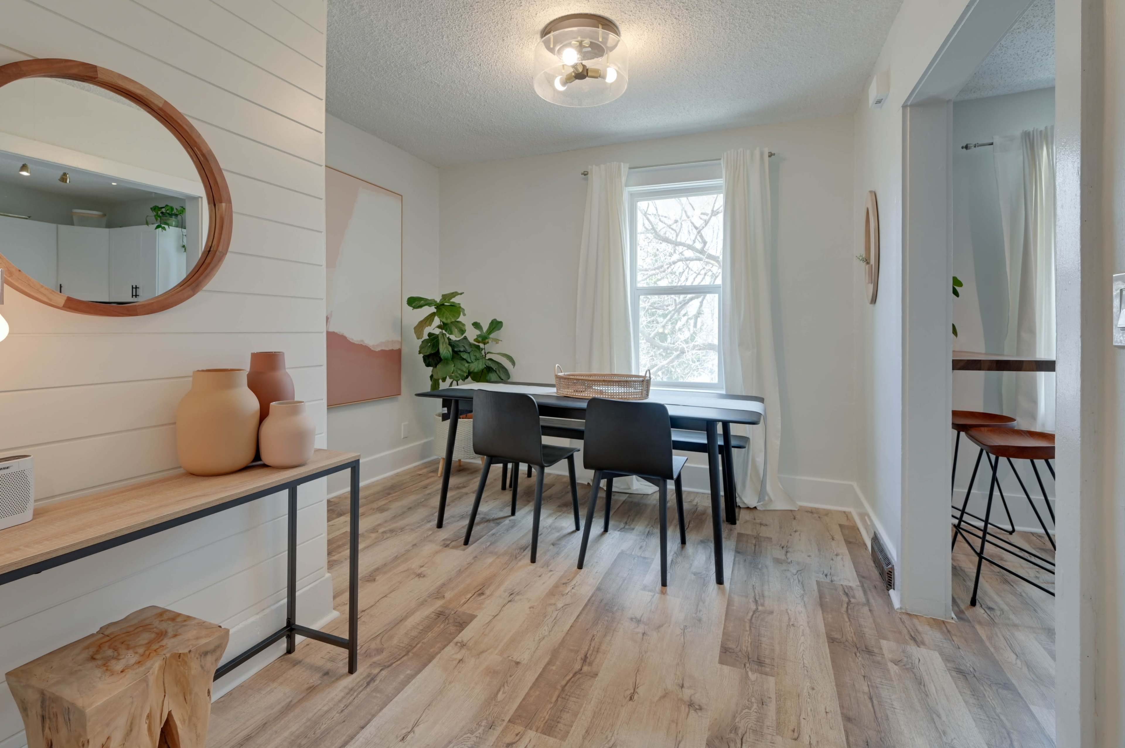 A bright dining area with a wooden table, black chairs, and a plant beside a window, complemented by a decorative mirror and vases on a console table.