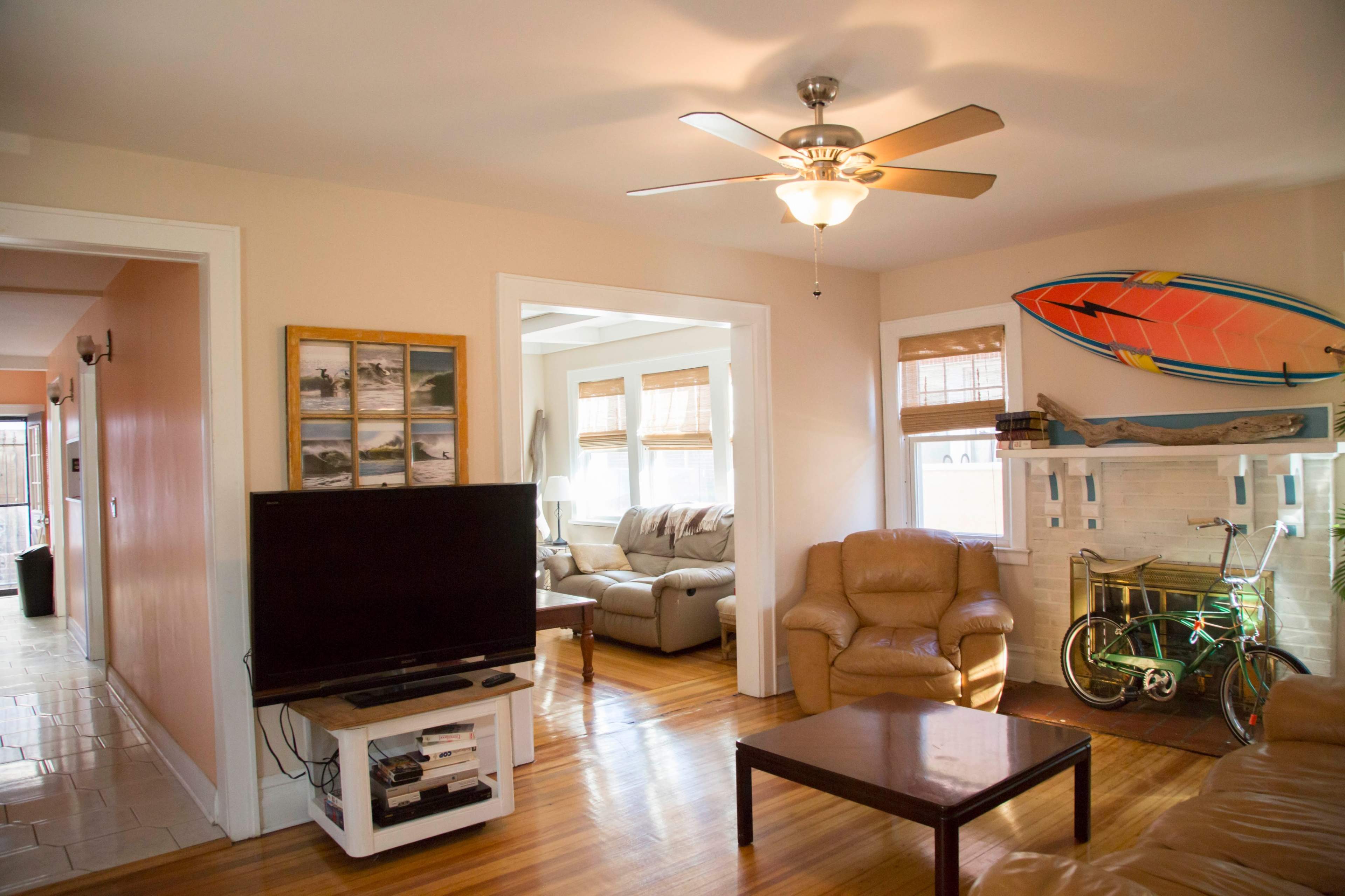A living room features a ceiling fan, a television on a white stand, a brown leather chair, a coffee table, and a bicycle next to a fireplace with a decorative surfboard on the wall.