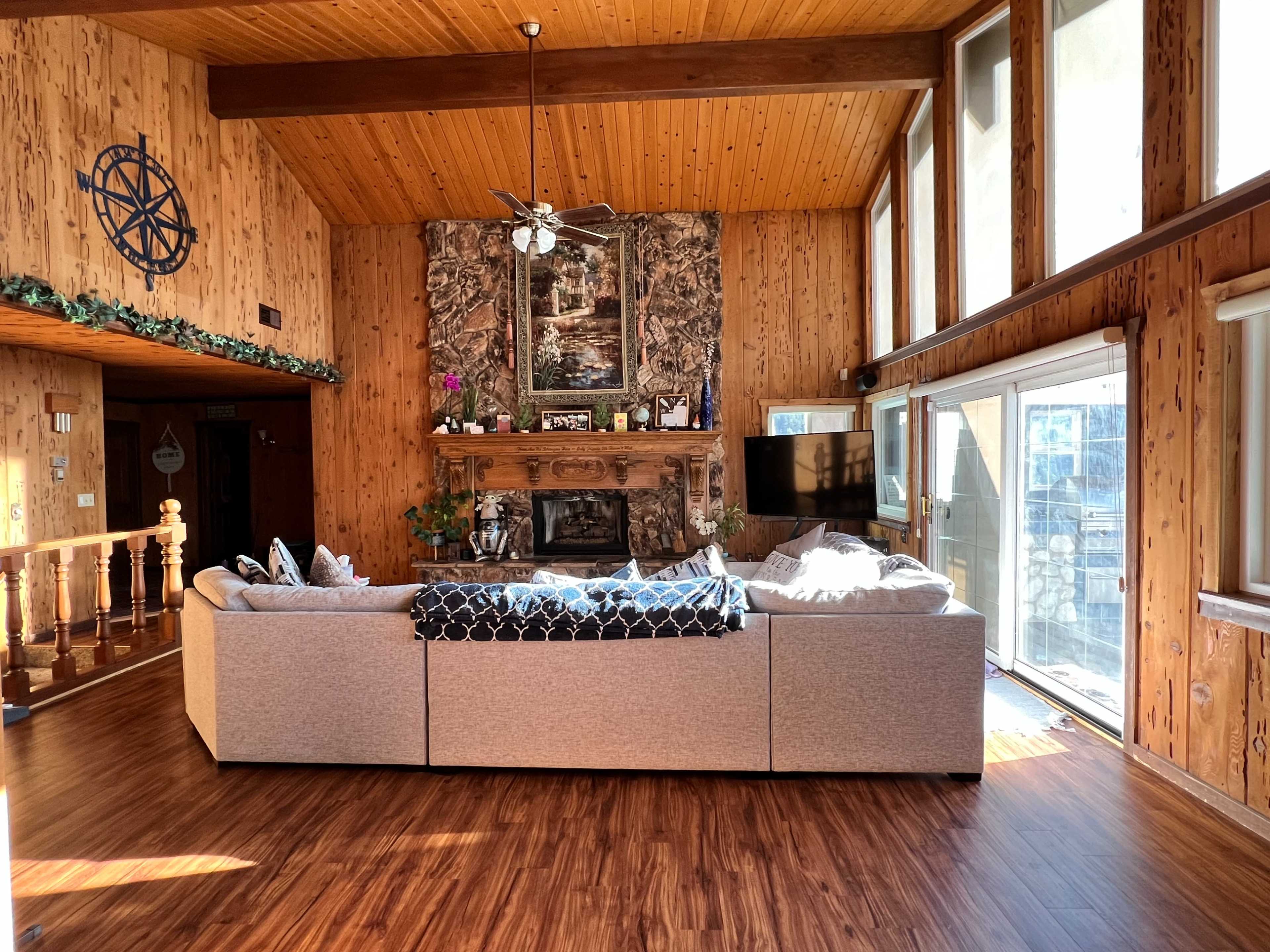 A spacious living room with wooden walls, a large stone fireplace, and a sectional sofa facing a wall of windows.