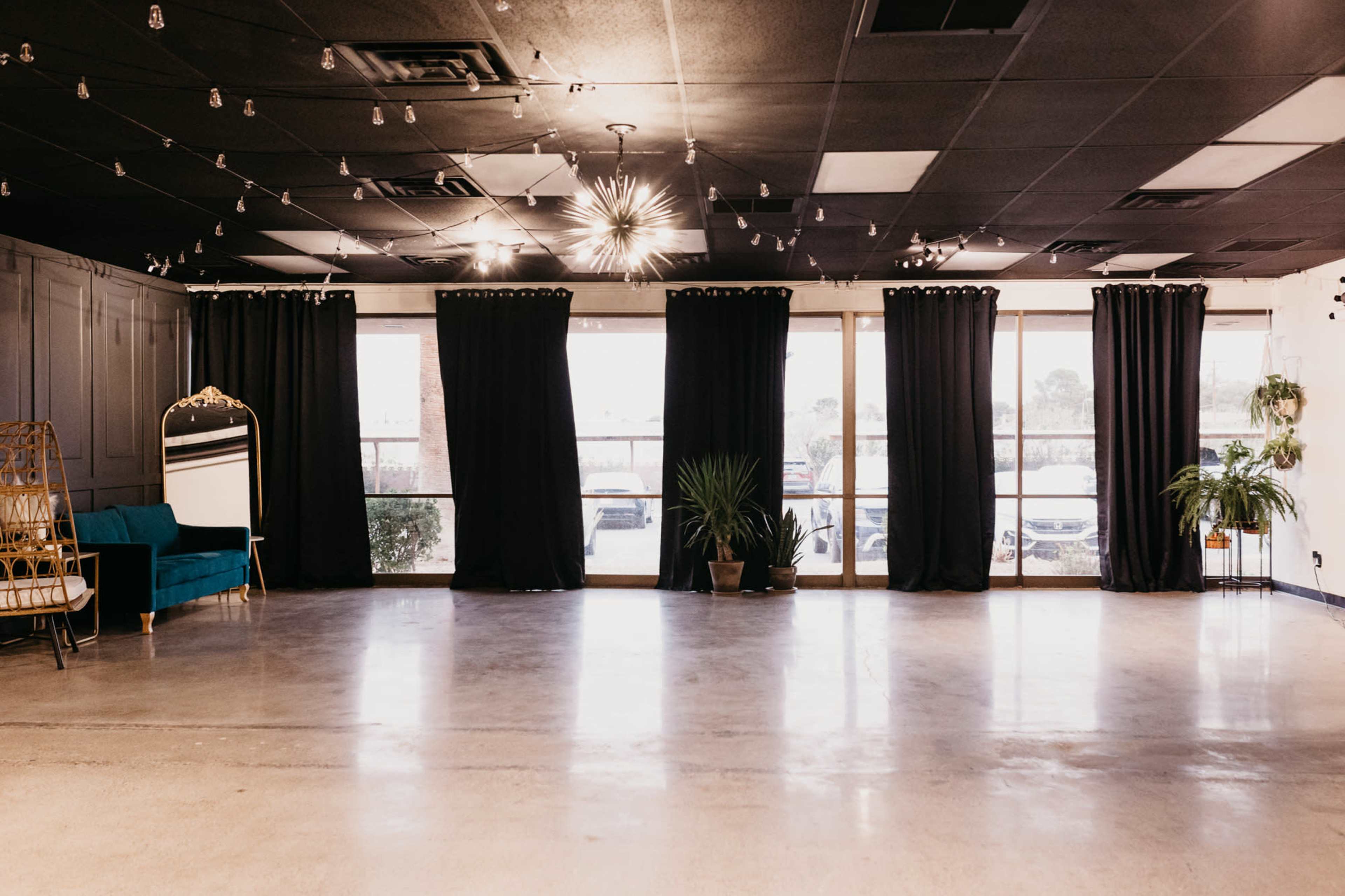 An empty event space with black curtains covering large windows, a teal couch against a wall, and potted plants in the corners.