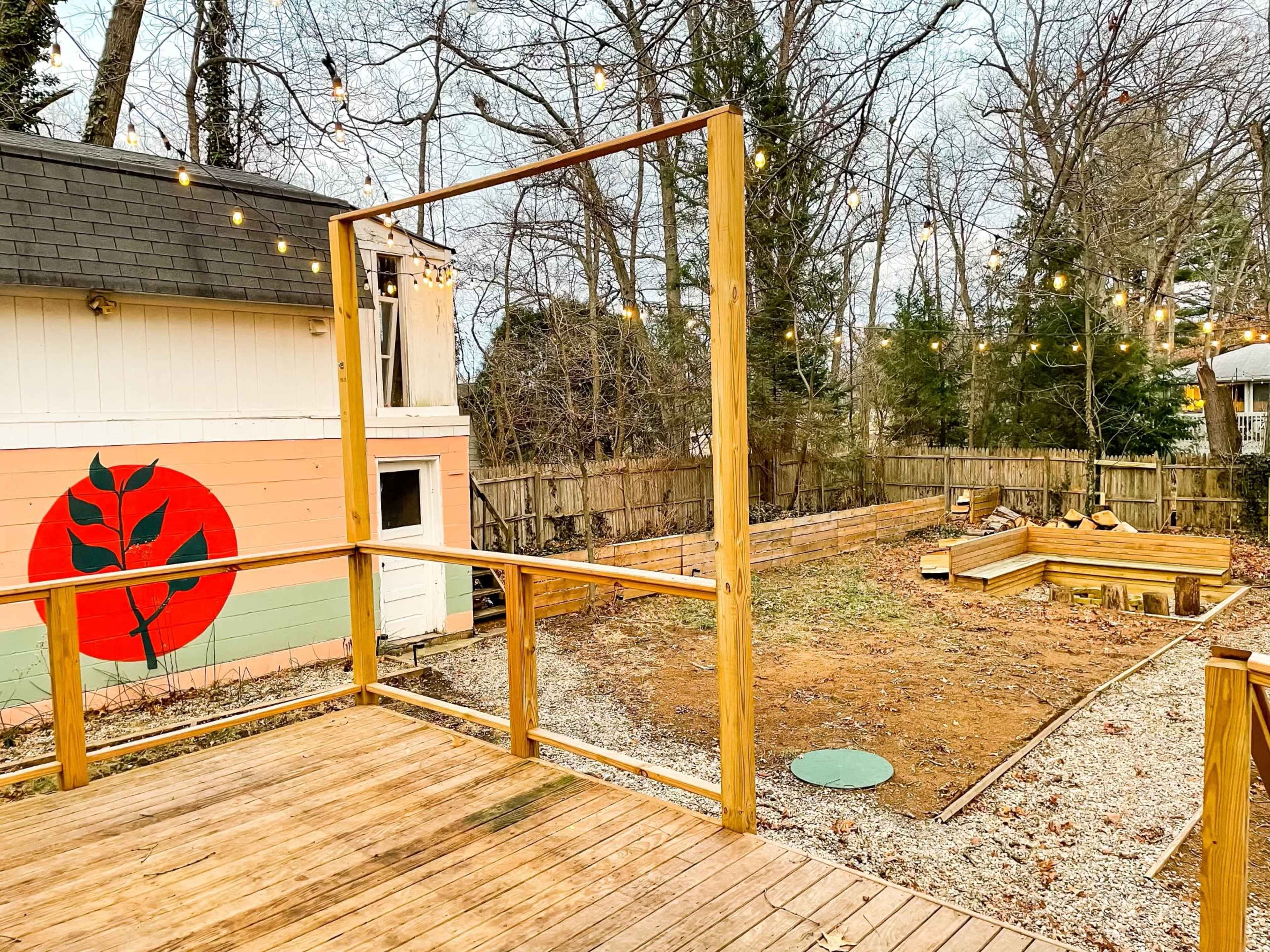 A wooden deck frames a view of a backyard with a circular garden bed, string lights overhead, and a house adorned with a mural featuring a large plant design.