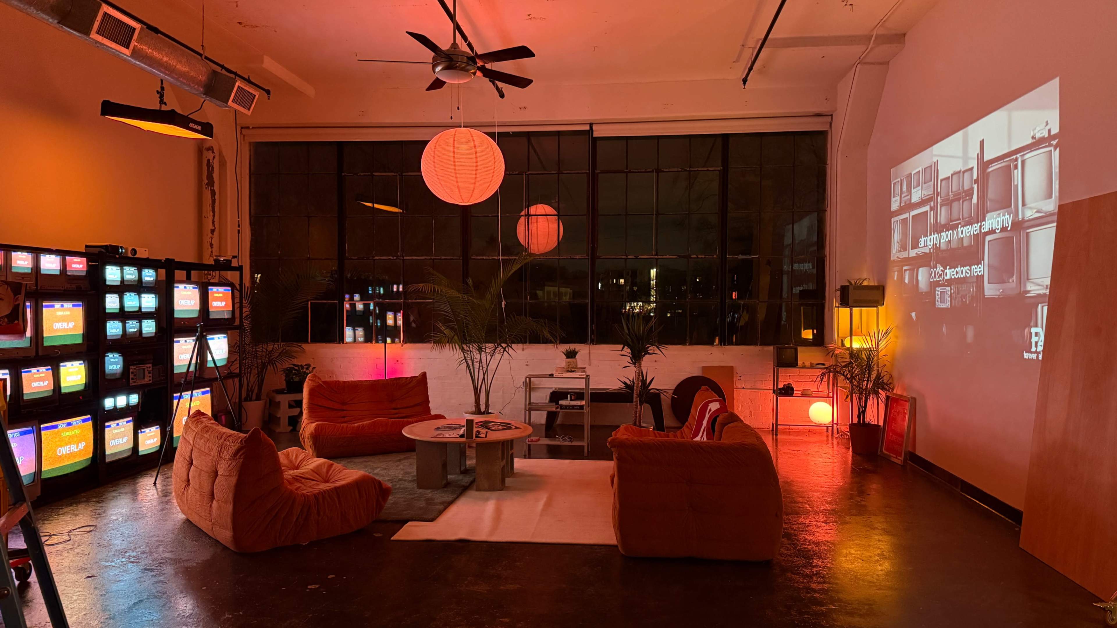 The scene shows a modern, low-light lounge with bright orange furniture, multiple retro televisions, and a large wall projection, creating a cozy ambiance.