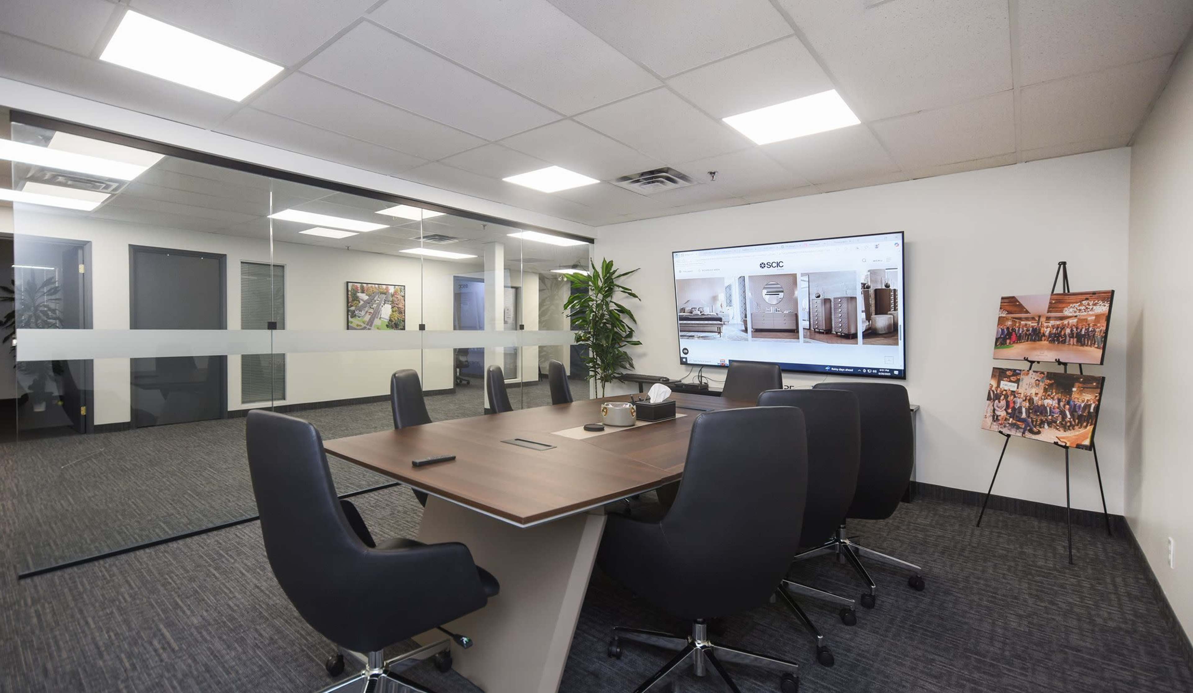 A modern conference room features a large table surrounded by ergonomic chairs and a display screen on the wall, with a glass partition separating it from an adjacent space.