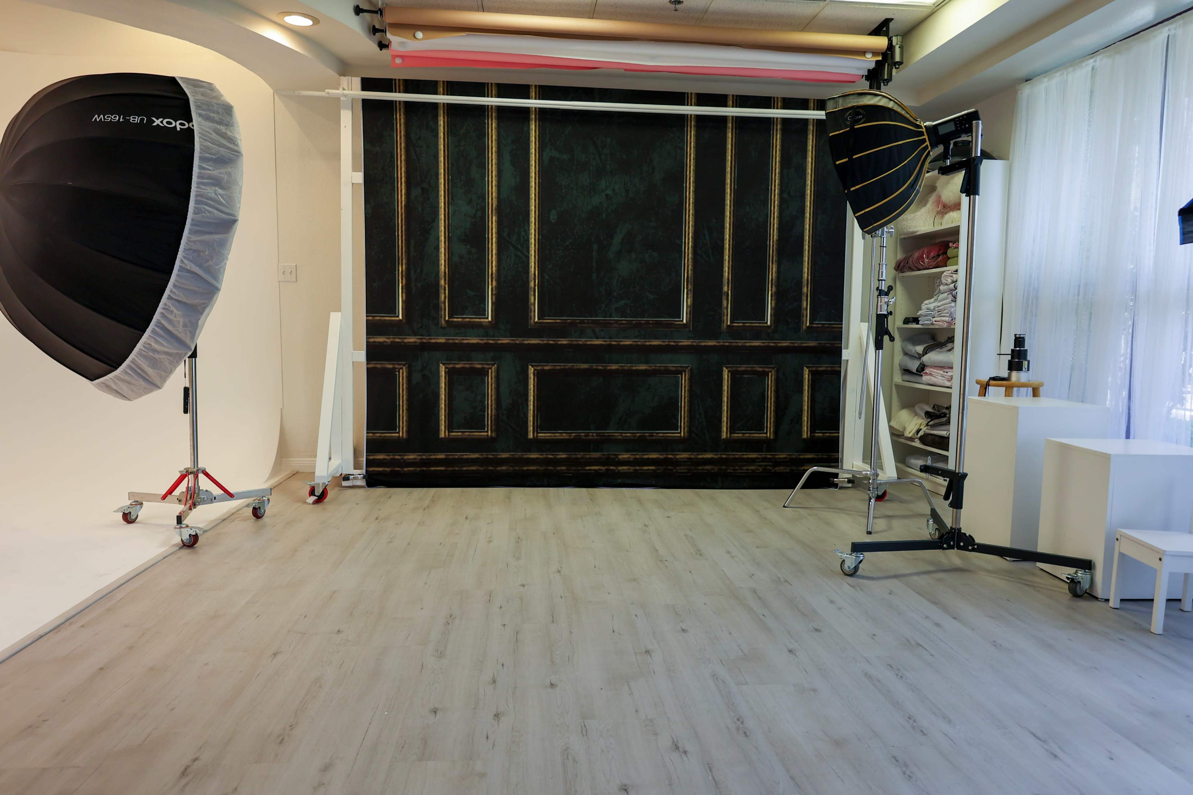 The image shows a photography studio with a dark-painted backdrop, bright lighting equipment, and wooden flooring.