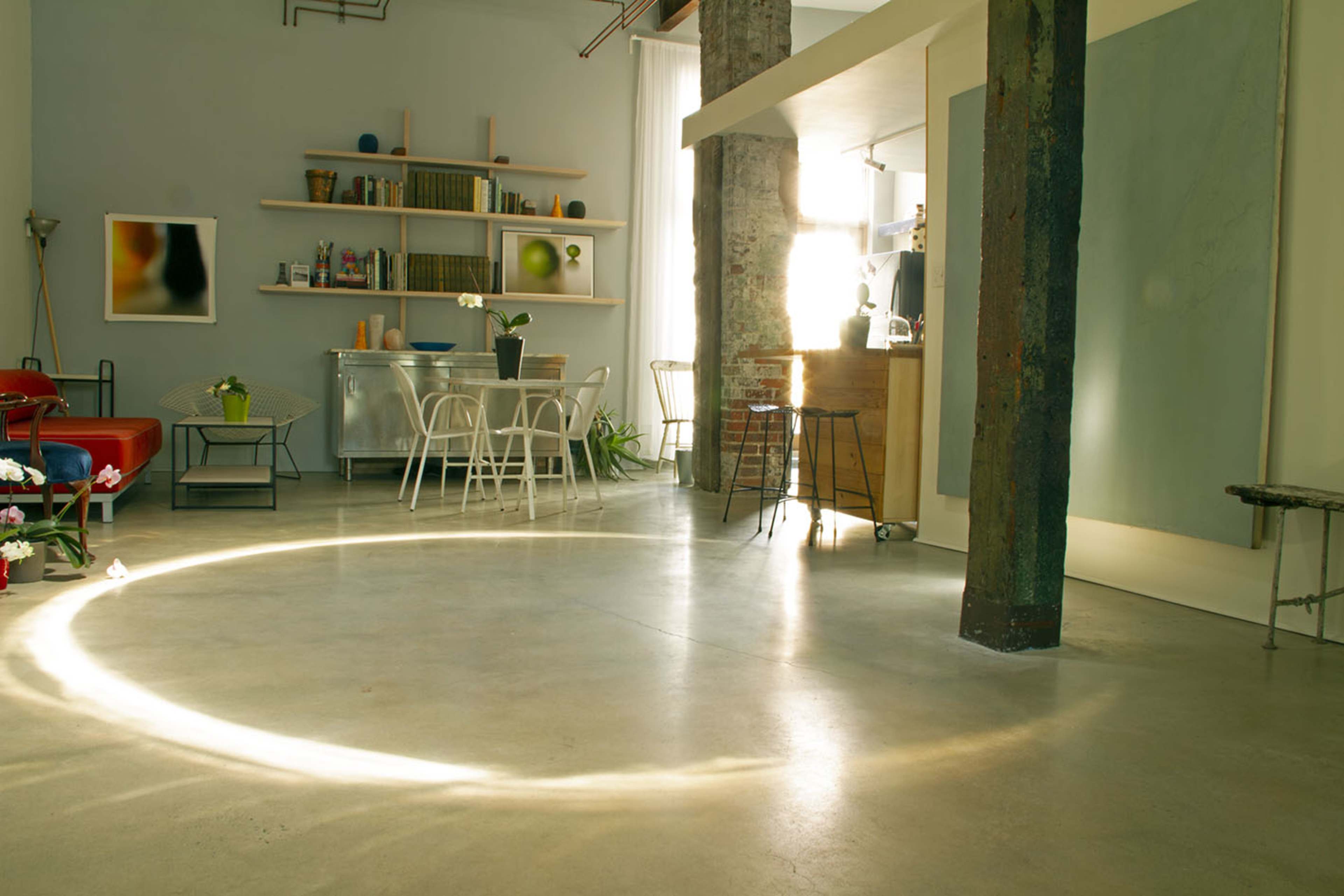 A modern, spacious room with concrete floors, featuring a sitting area with a red couch, a dining area with white chairs, and shelves filled with books and decor.