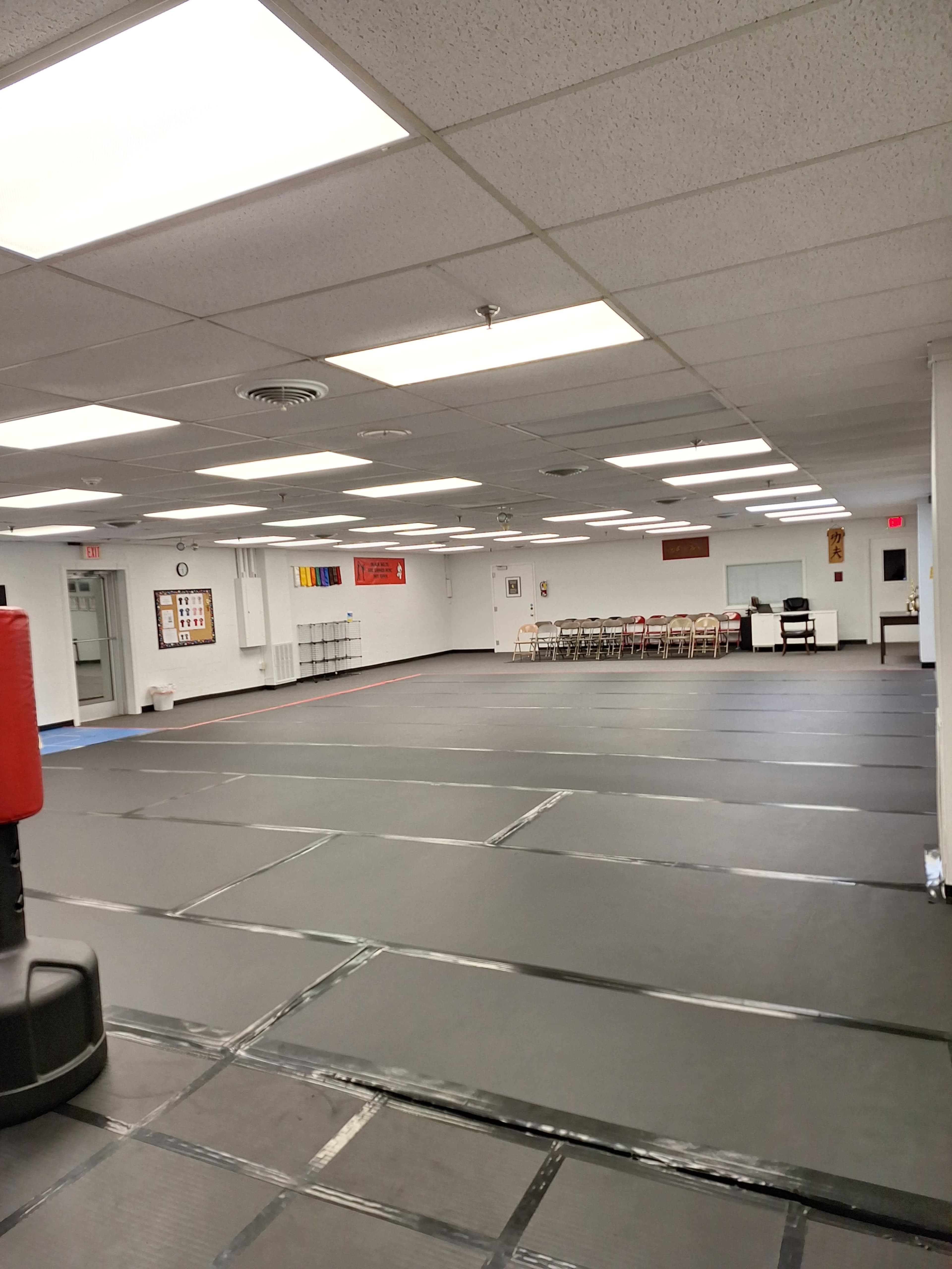 The image shows an empty martial arts studio with black mats on the floor and a few chairs along the walls.