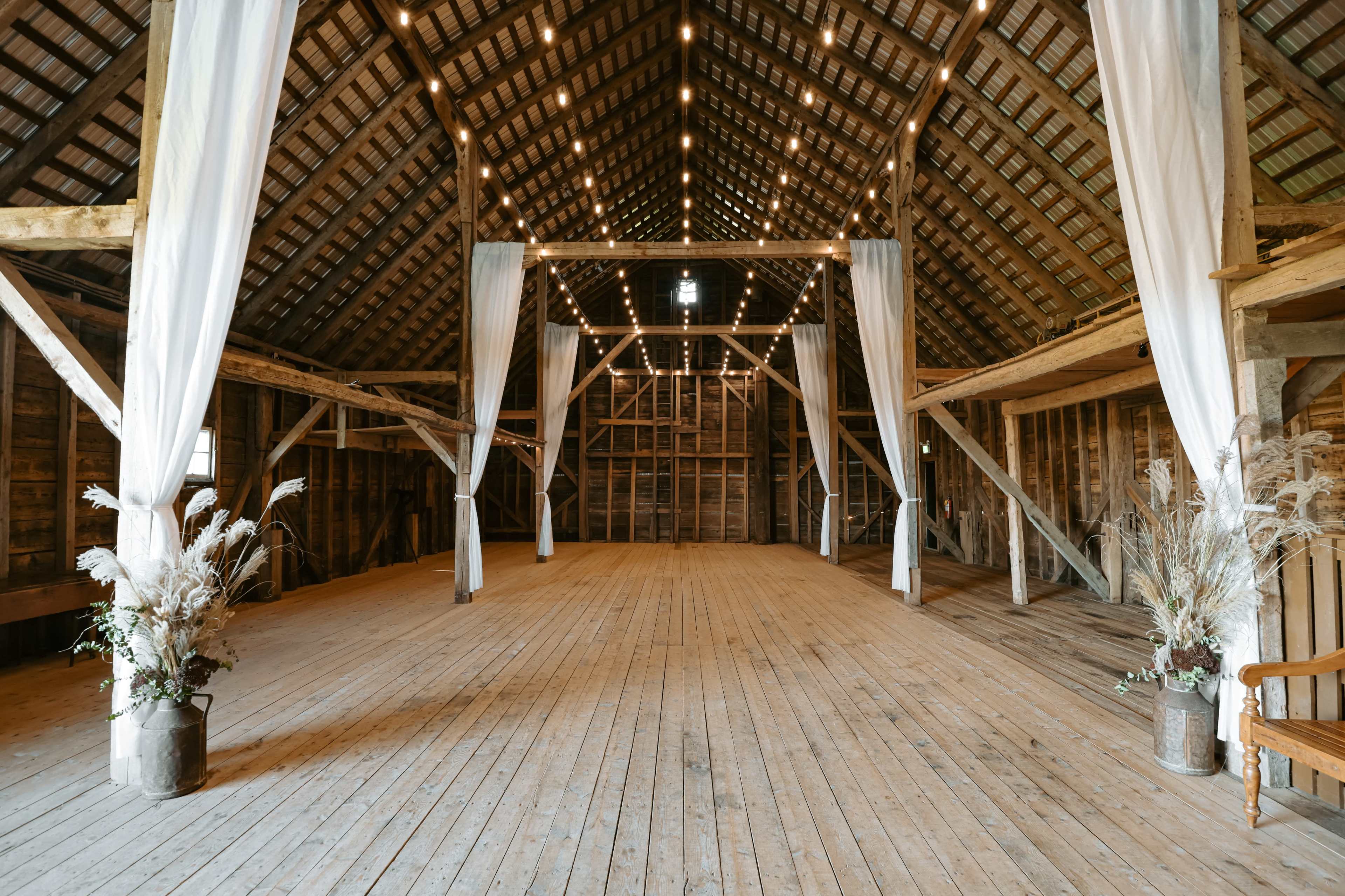 The interior of a spacious barn features high wooden beams, a wooden floor, and white drapery hanging from the ceiling, with flower arrangements placed in the corners.