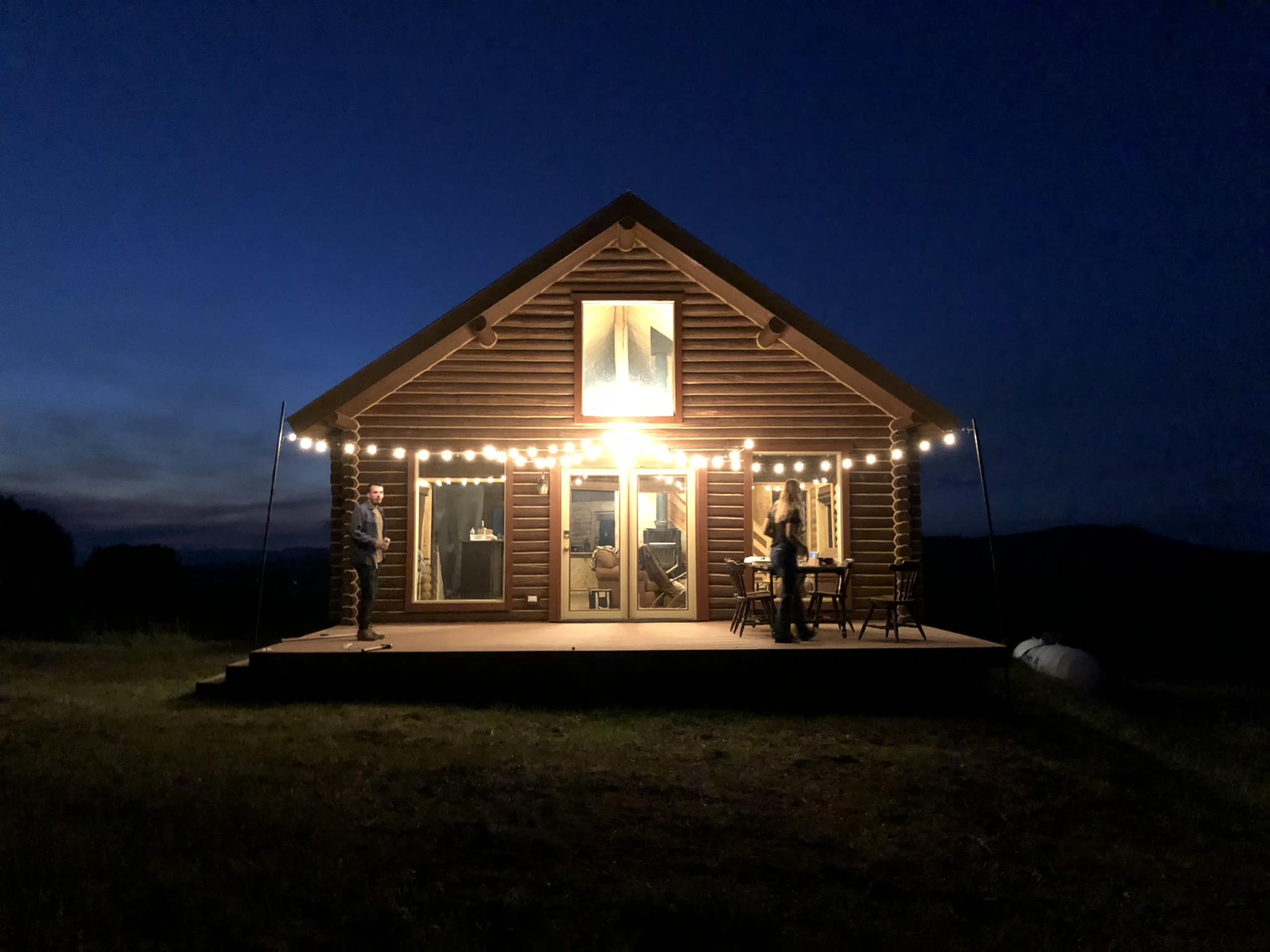 A wooden cabin is illuminated by string lights at night, with people gathered on the porch.