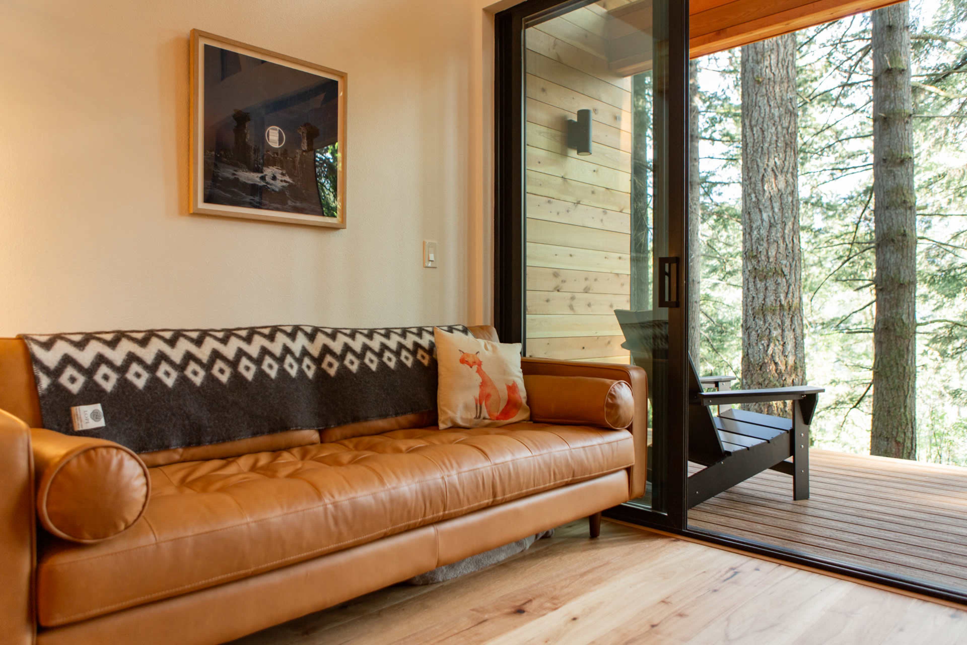 A leather sofa with a patterned throw pillow is positioned next to a large sliding glass door that opens to a wooden deck surrounded by trees.