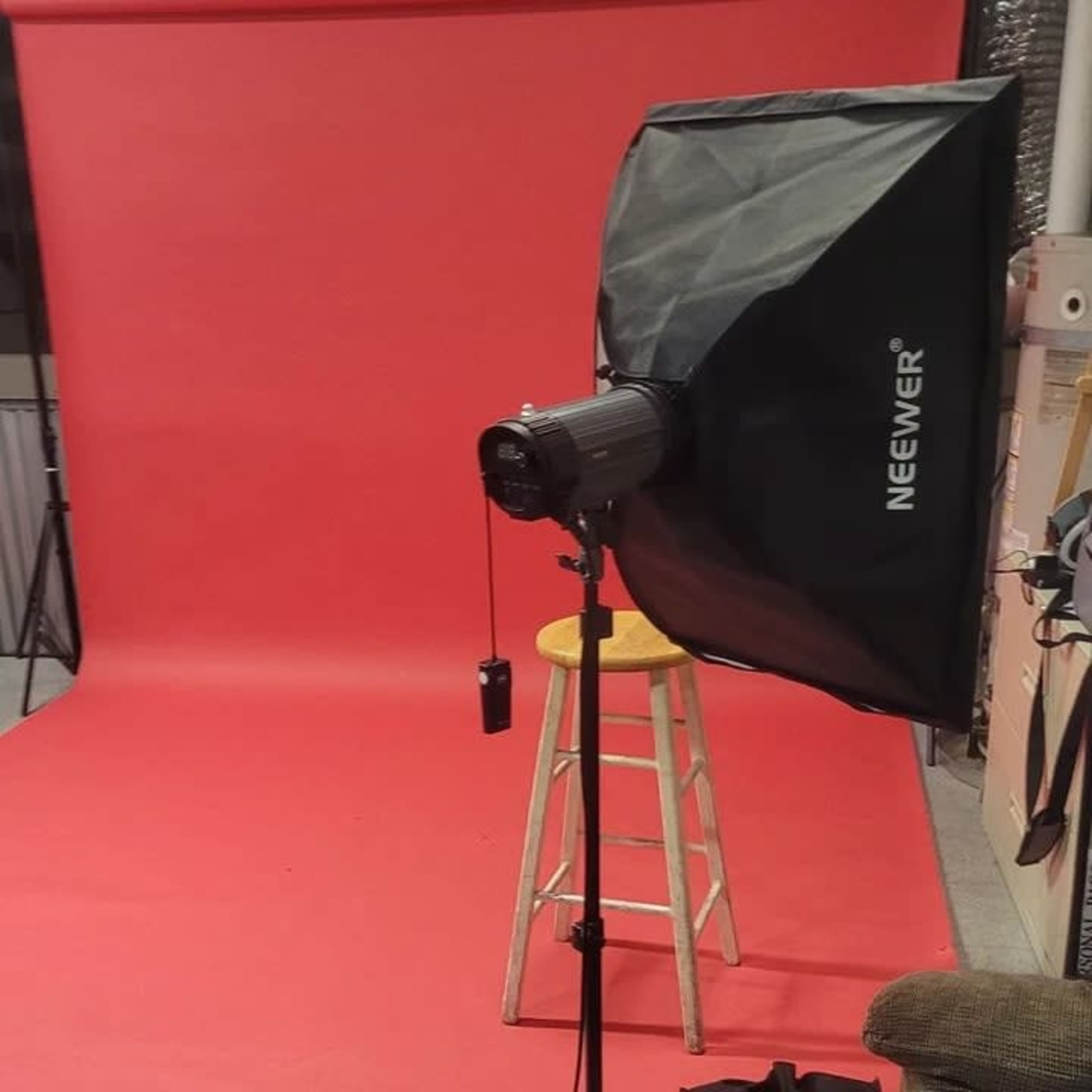 A studio setup features a red backdrop, a light stand with a softbox, and a wooden stool.