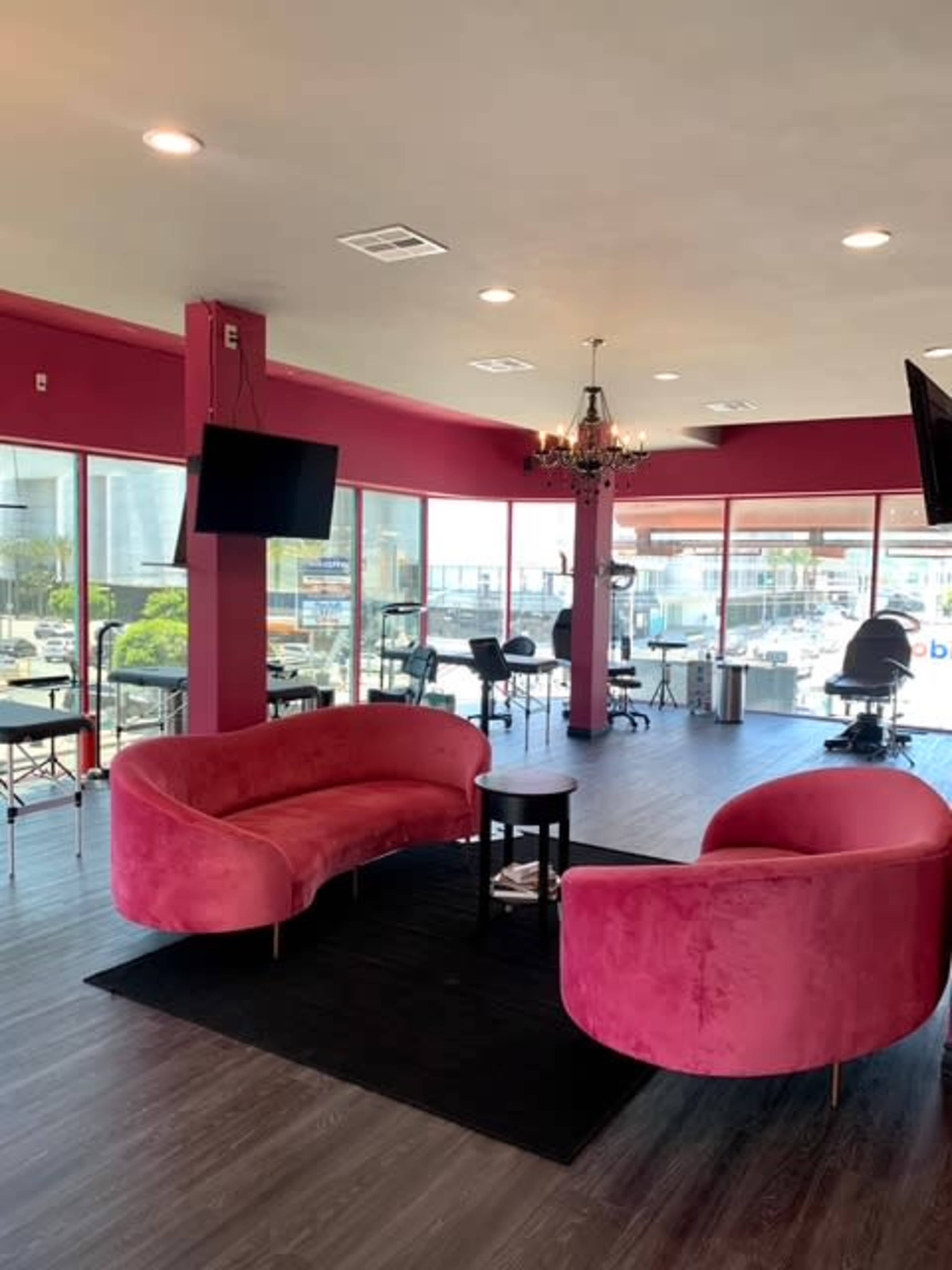 The image shows a bright salon interior with pink walls, two curved pink sofas, and a chandelier, along with salon stations visible through large windows.