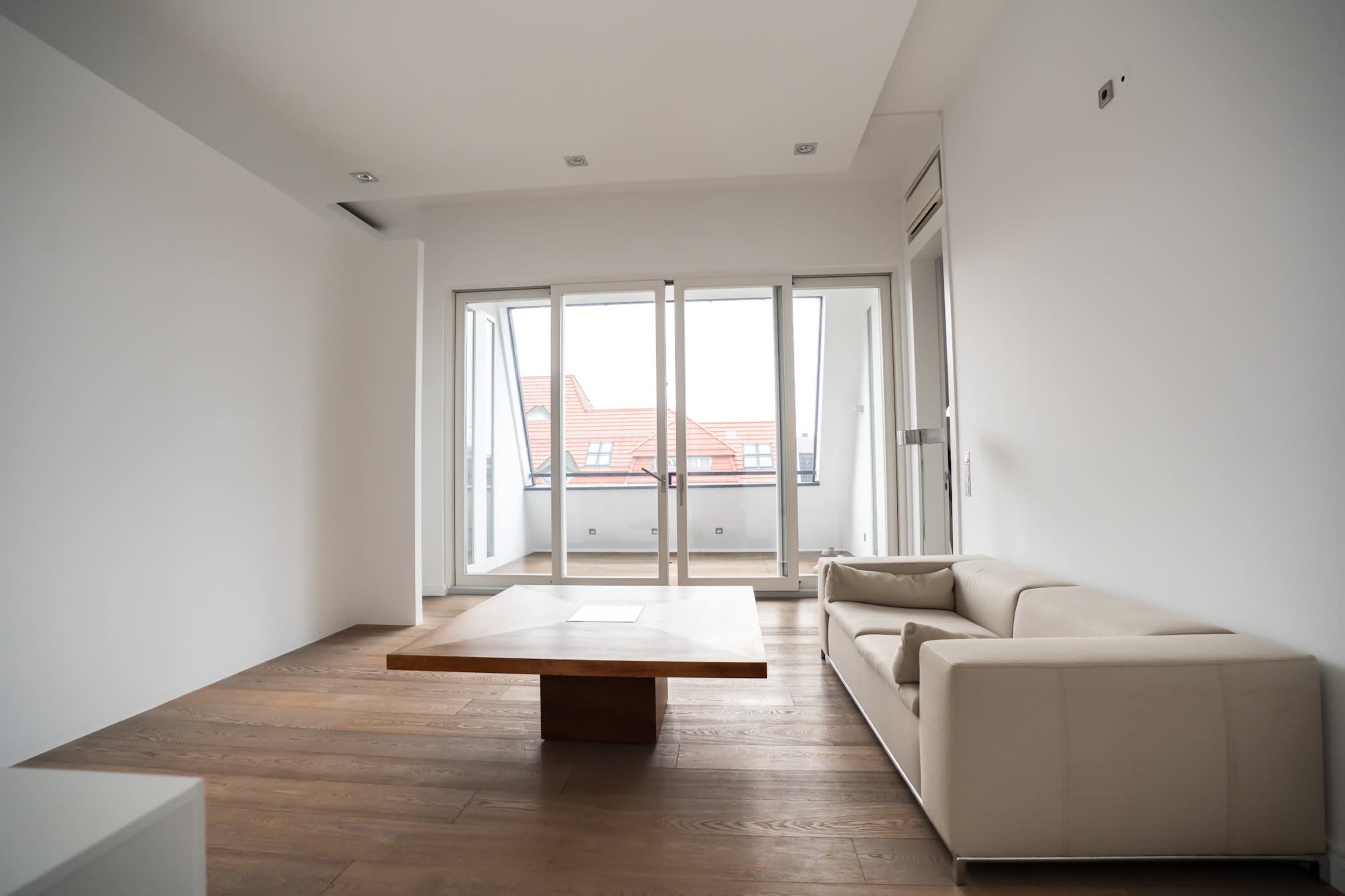 The image shows a minimalist living room with a large wooden table and a white sofa, featuring floor-to-ceiling windows that open to a balcony.