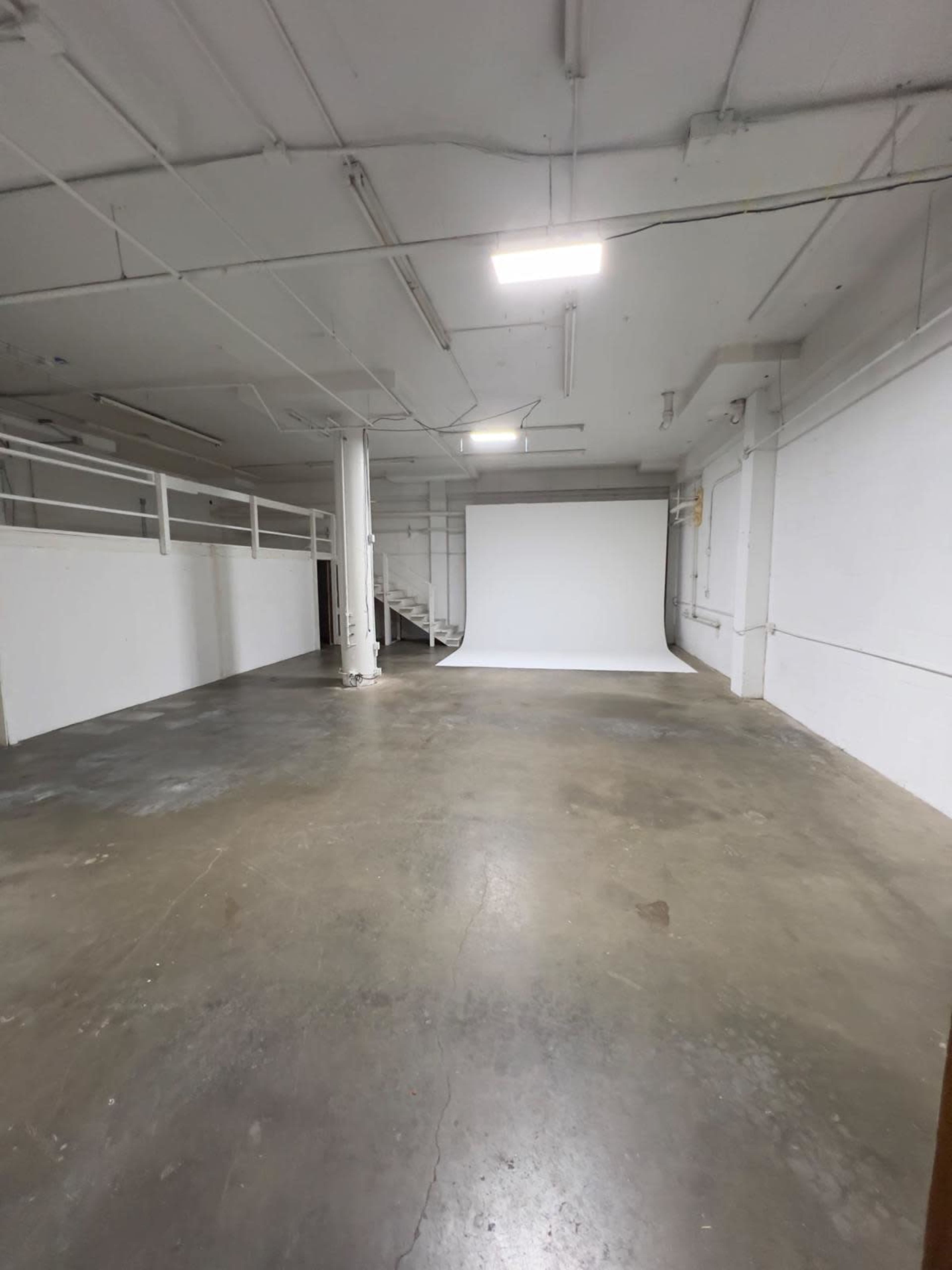 The image shows a spacious, empty industrial studio with a white backdrop at one end and exposed pipes on the ceiling.