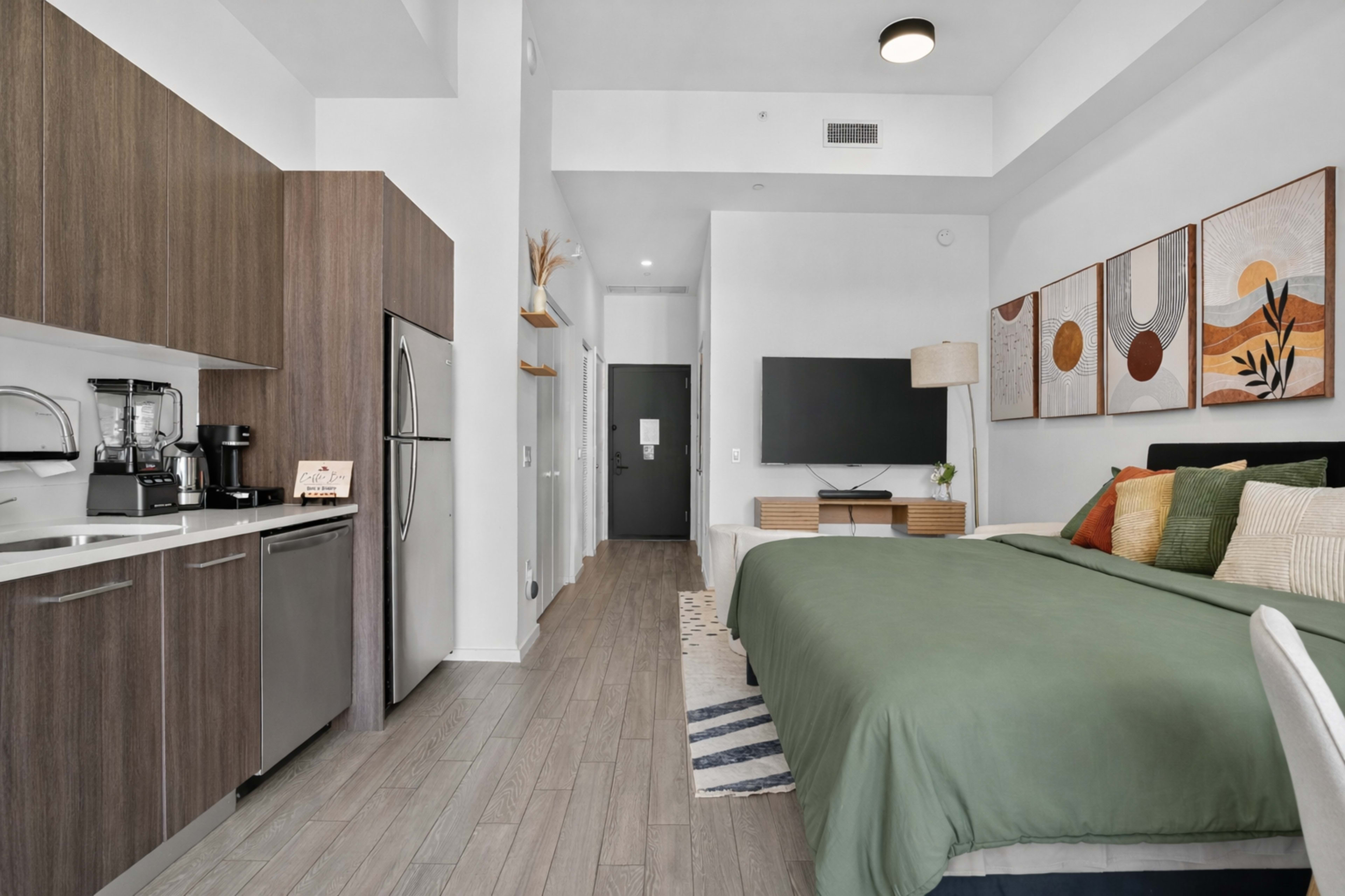 The image shows a modern studio apartment featuring a kitchenette, a bed with a green comforter, and decorative wall art.