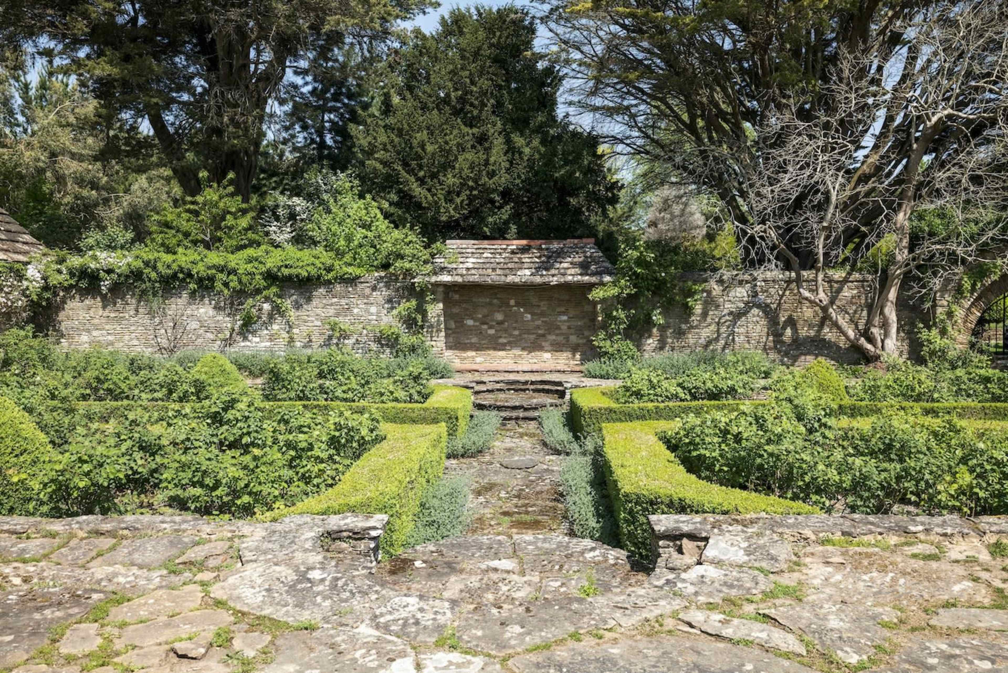 The image shows a well-maintained garden with stone pathways, bordered by neatly trimmed hedges and a backdrop of greenery behind a stone wall.