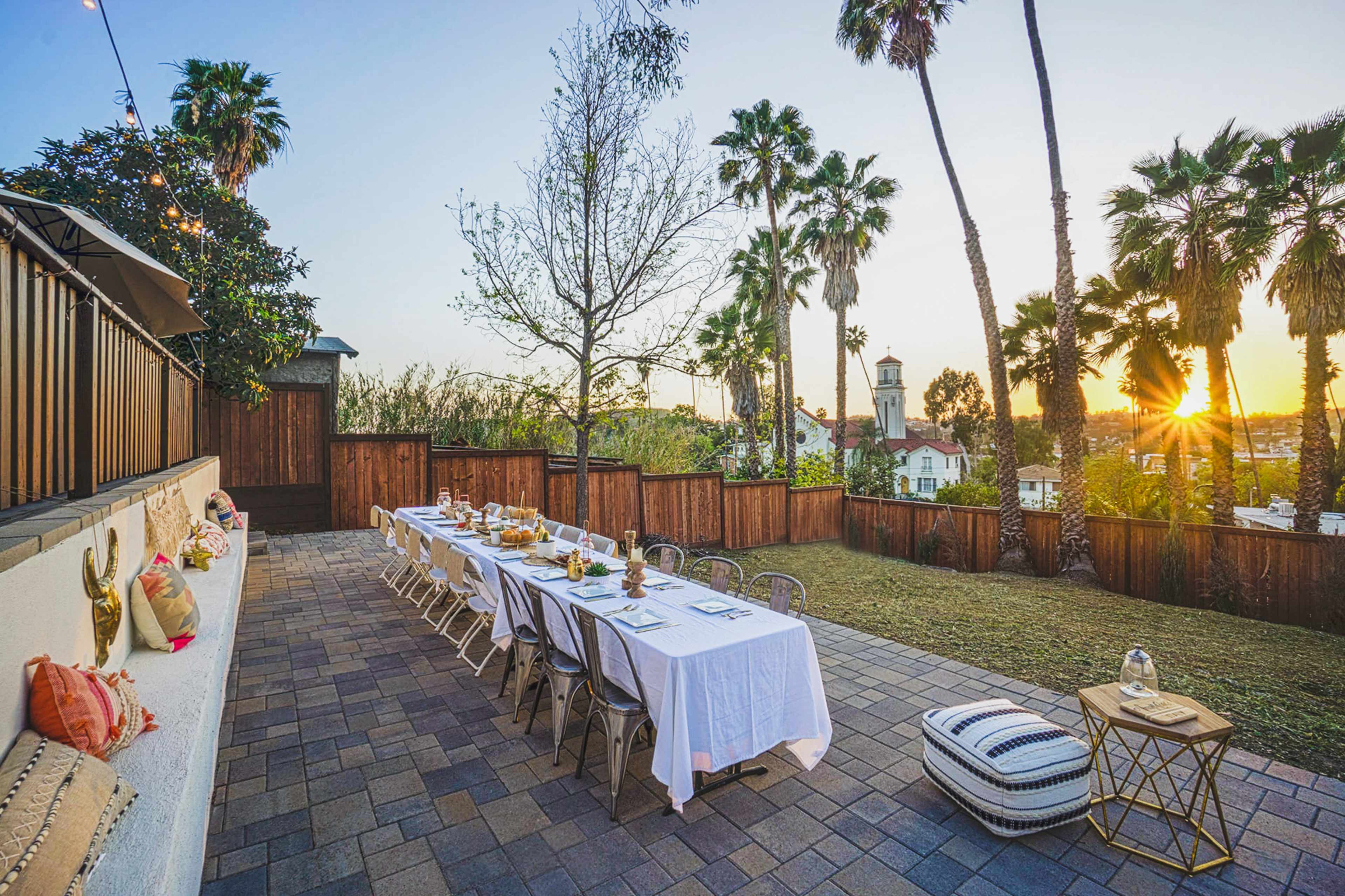 A long dining table is set outdoors under palm trees, with a sunset in the background illuminating the scene.