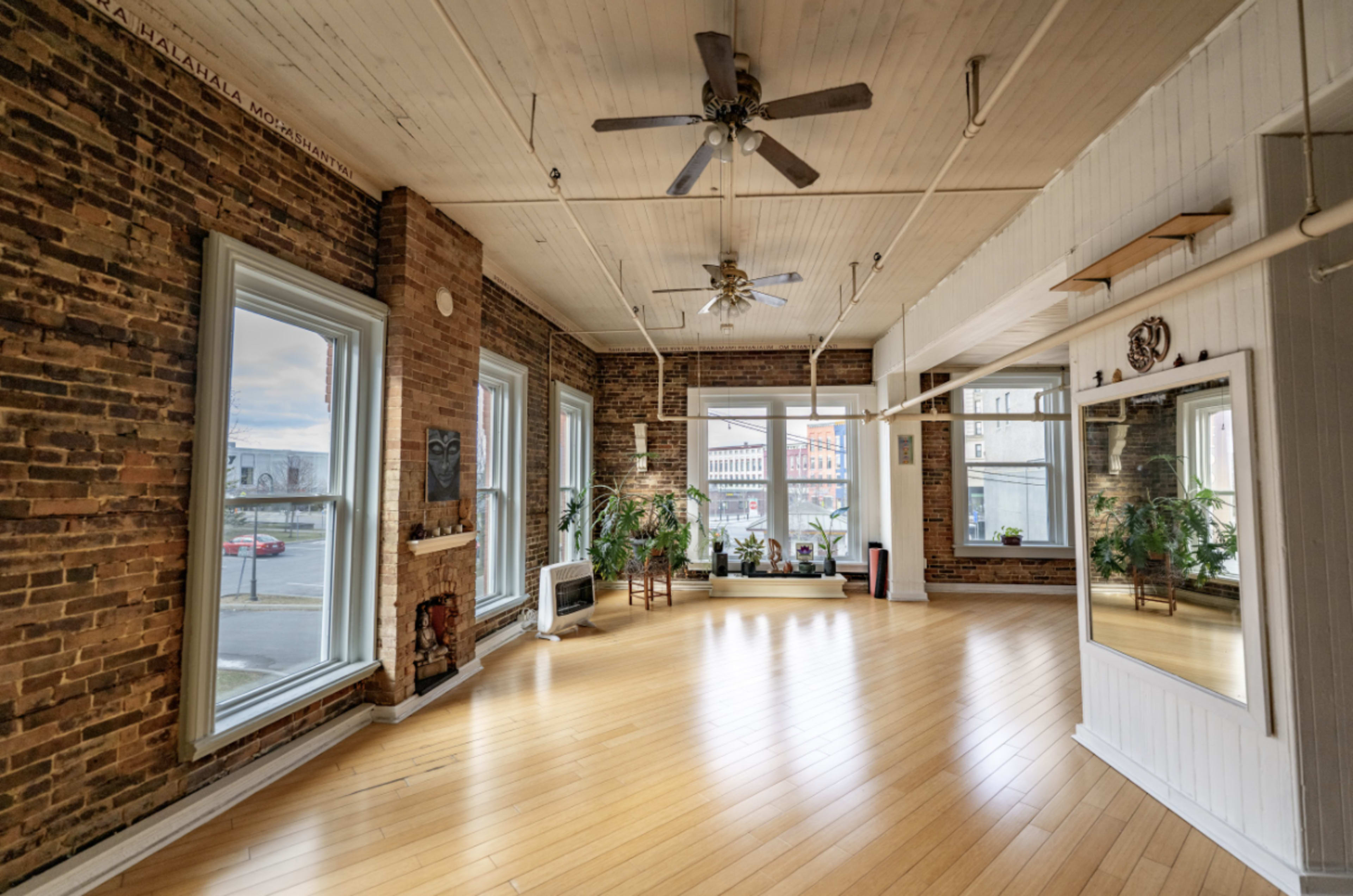 The image shows a spacious room with exposed brick walls, large windows, wooden floors, and plants, featuring a mirror and ceiling fans.