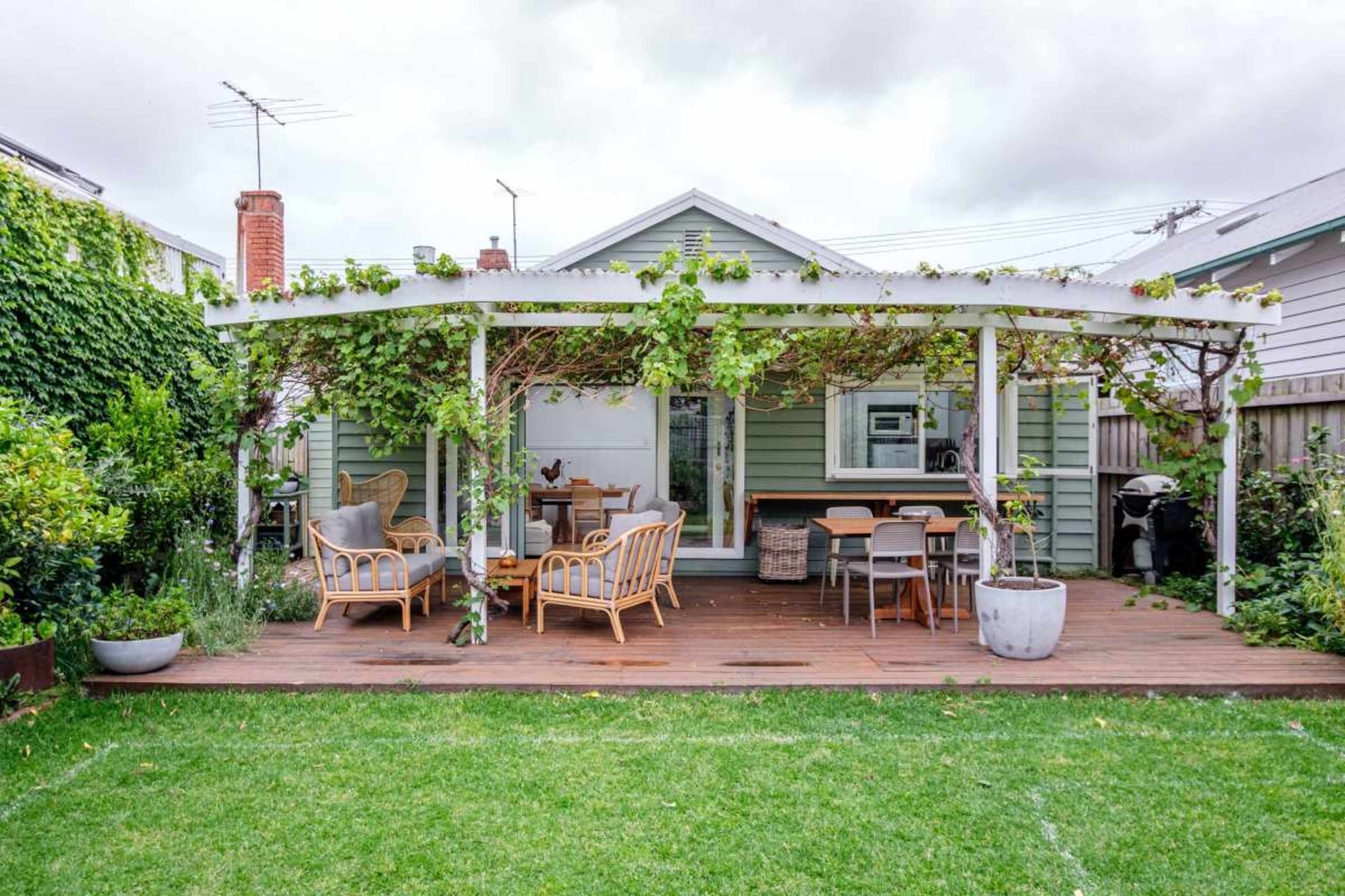 A wooden deck with seating and a dining area is covered by a pergola adorned with greenery, leading to a modest house.