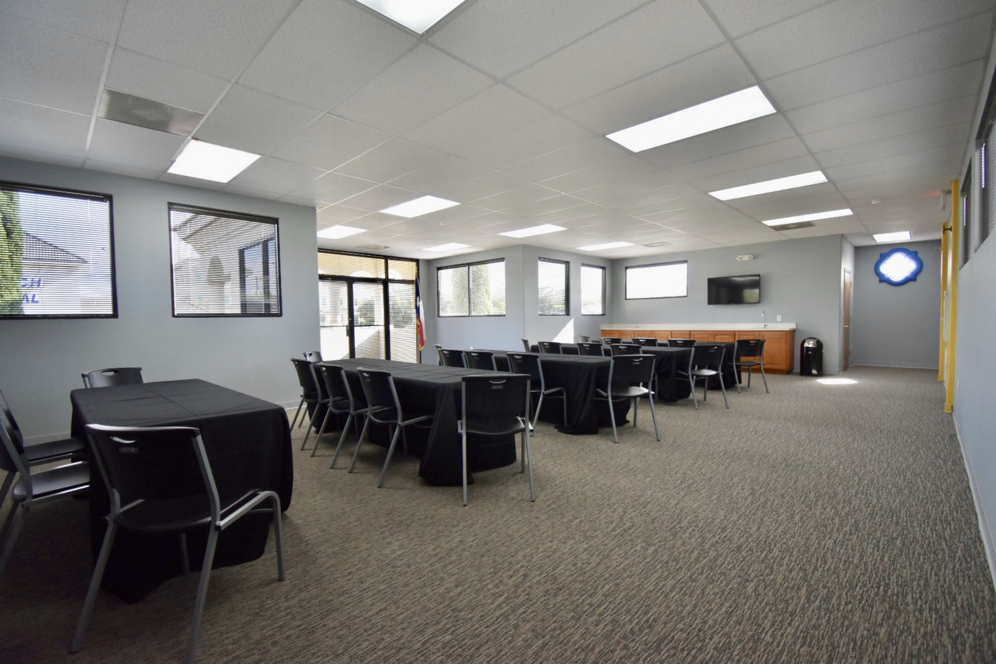 A conference room features multiple tables covered in black cloth, with chairs arranged neatly and large windows allowing natural light in.