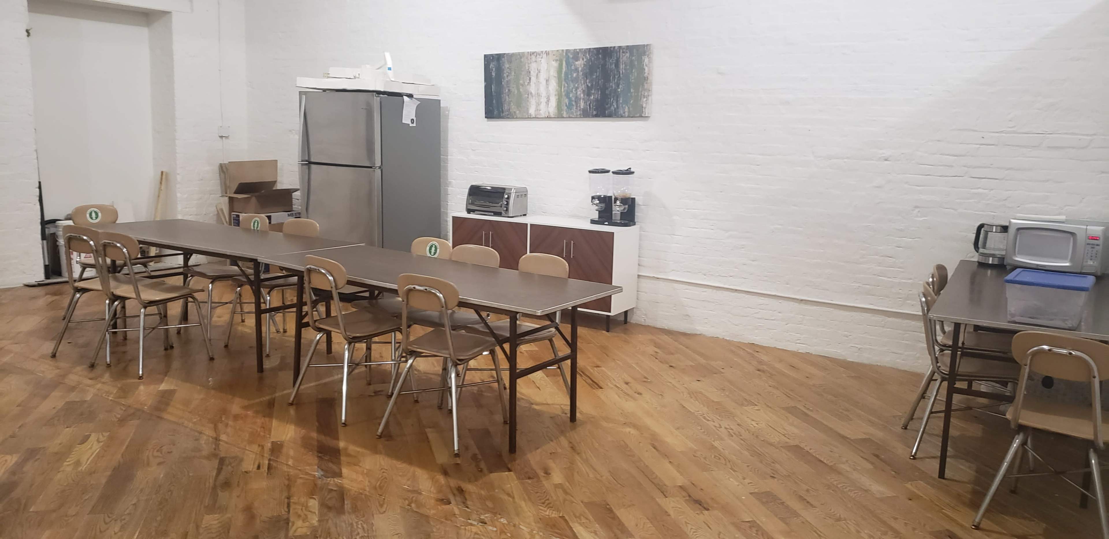 The image shows a spacious room with wooden floors, featuring a long table surrounded by chairs, a kitchen area with a refrigerator, microwave, and coffee maker, and a decorative artwork on the wall.