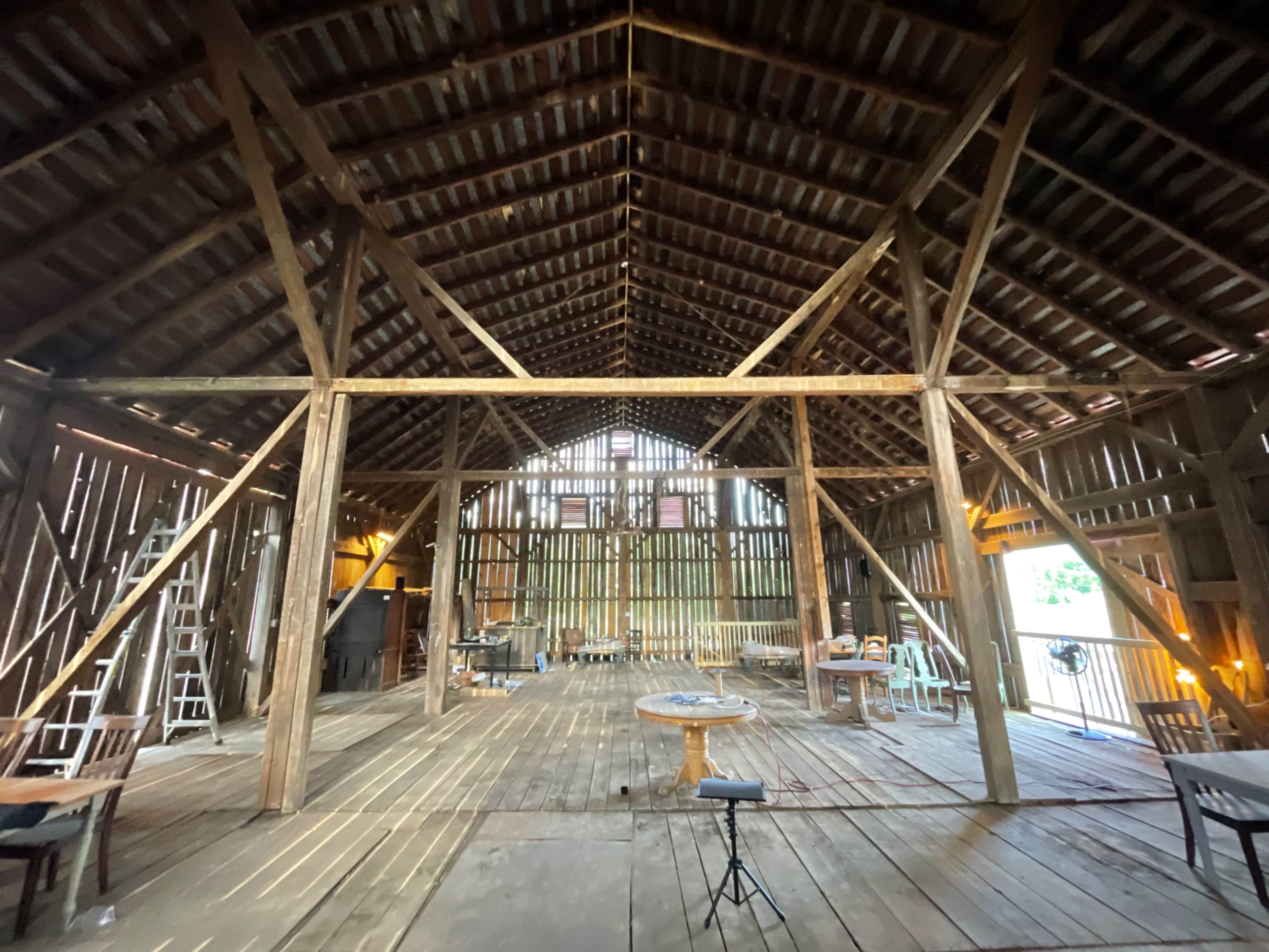 The interior of a spacious, rustic barn with exposed wooden beams, various furniture pieces, and tools scattered across the wooden floor.