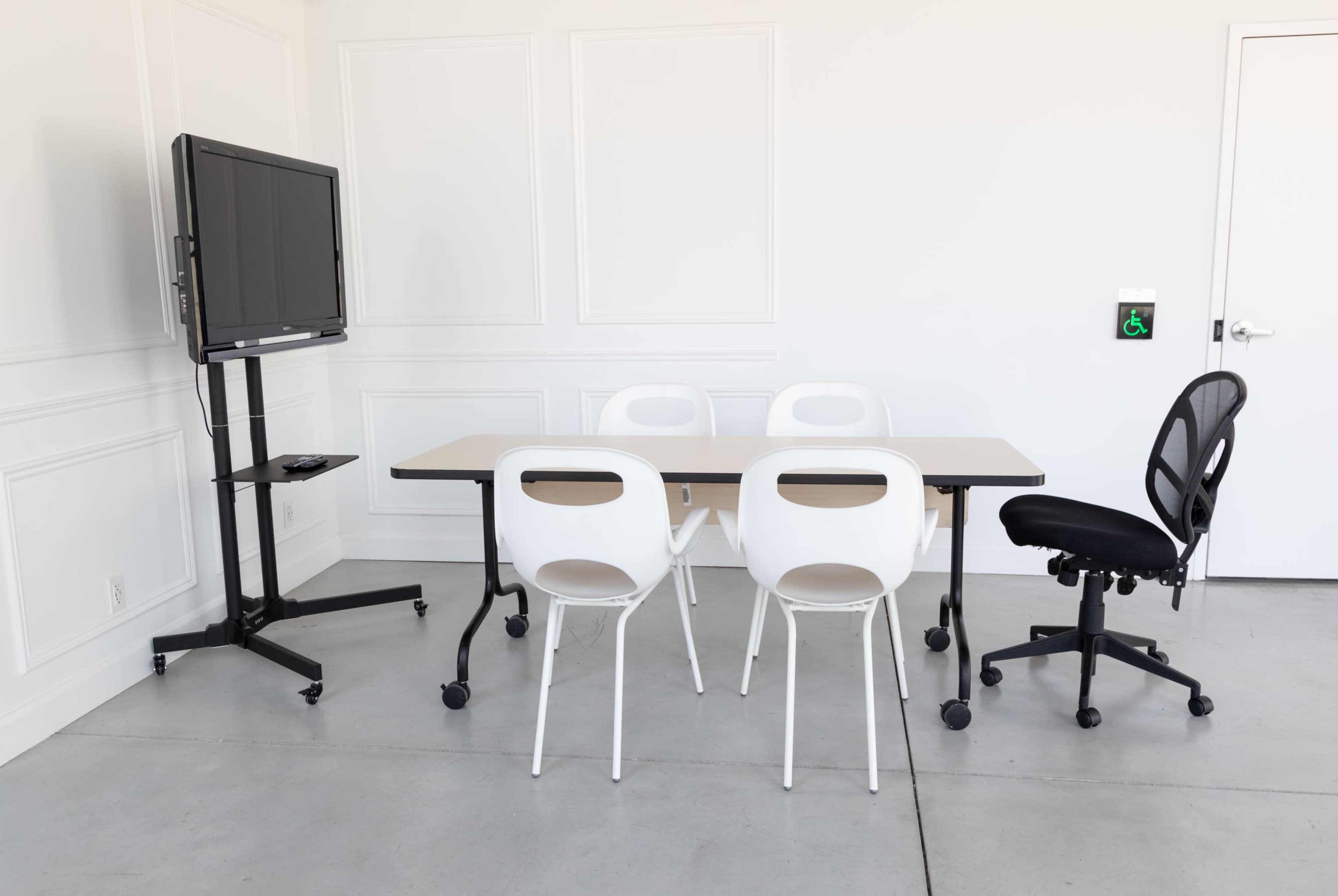 A meeting room features a rectangular table surrounded by four white chairs, with a black rolling office chair and a mounted television on a stand.