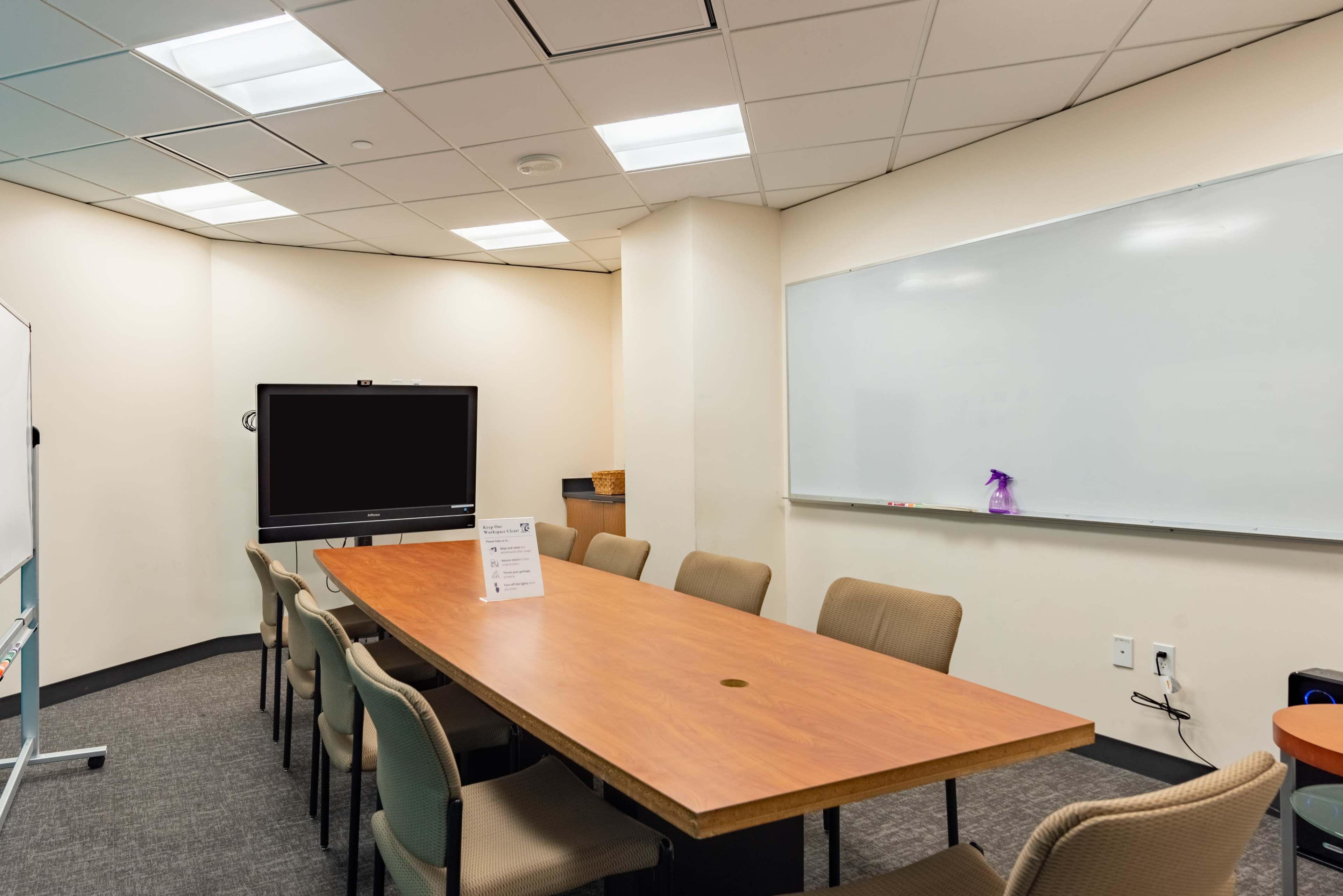 A conference room features a long wooden table surrounded by beige chairs, a large screen on one wall, and a whiteboard on another.