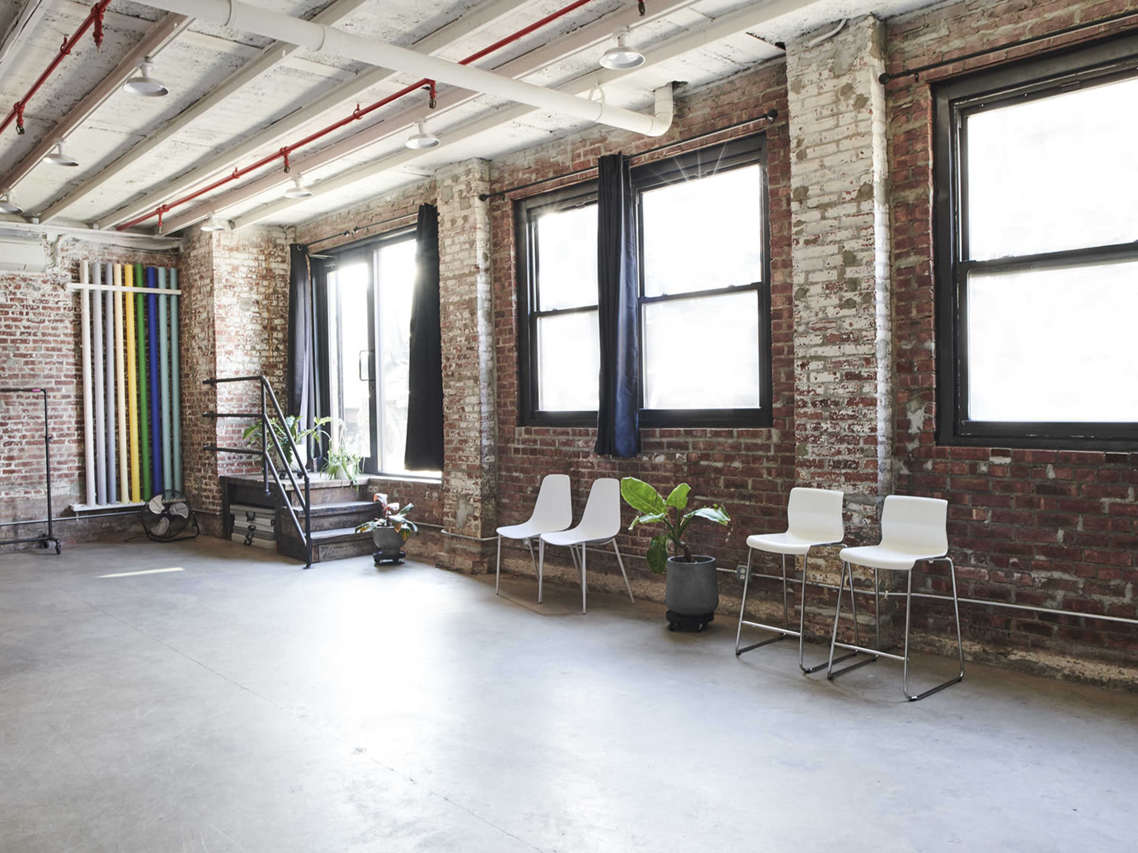 An interior space features exposed brick walls, large windows with black curtains, and three white chairs arranged on a concrete floor.