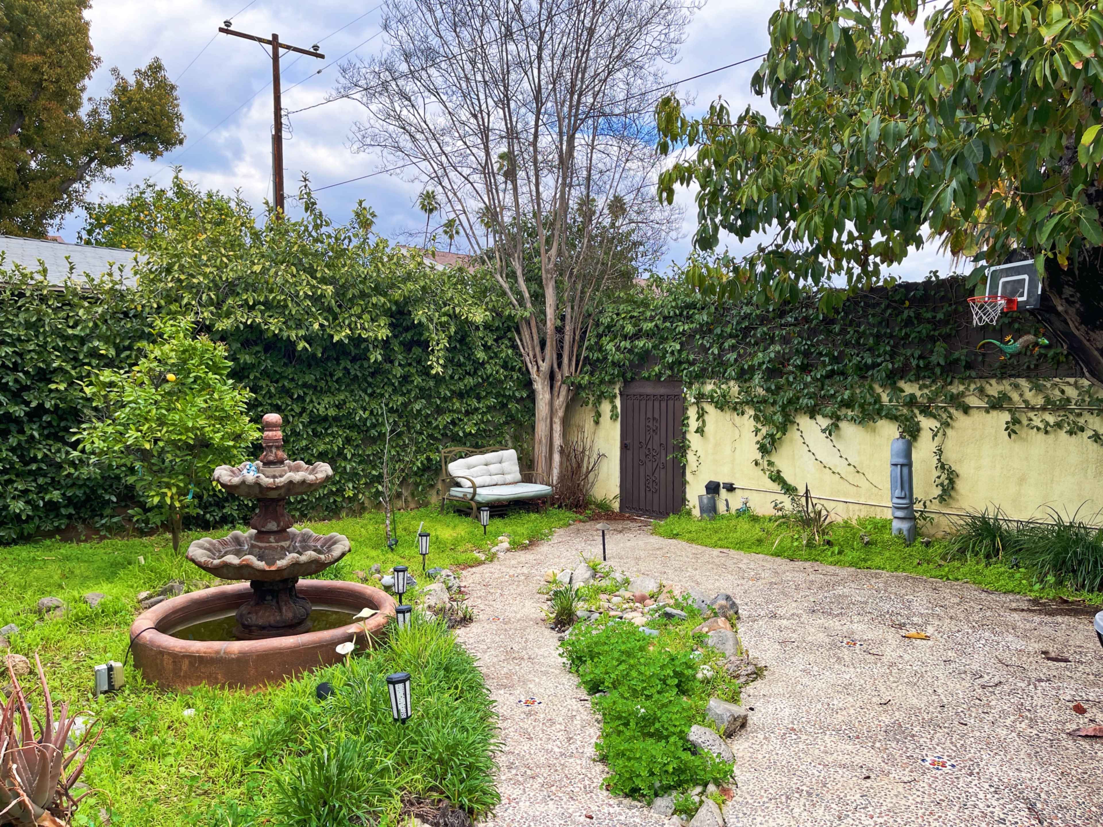 A small garden features a fountain, a bench, and surrounding greenery with a gravel path.