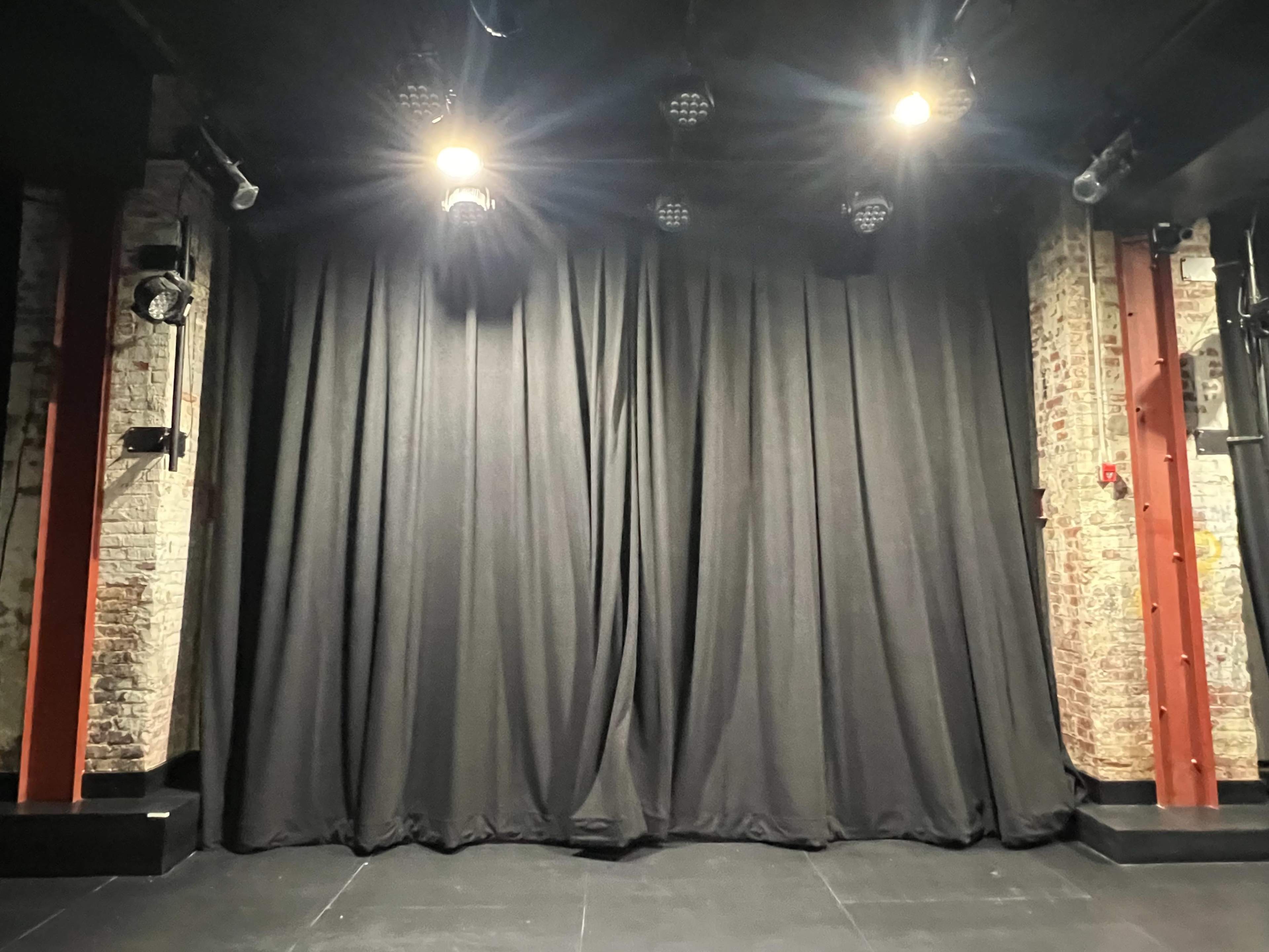 The image shows a dark stage with a black curtain in front, illuminated by several spotlights above.
