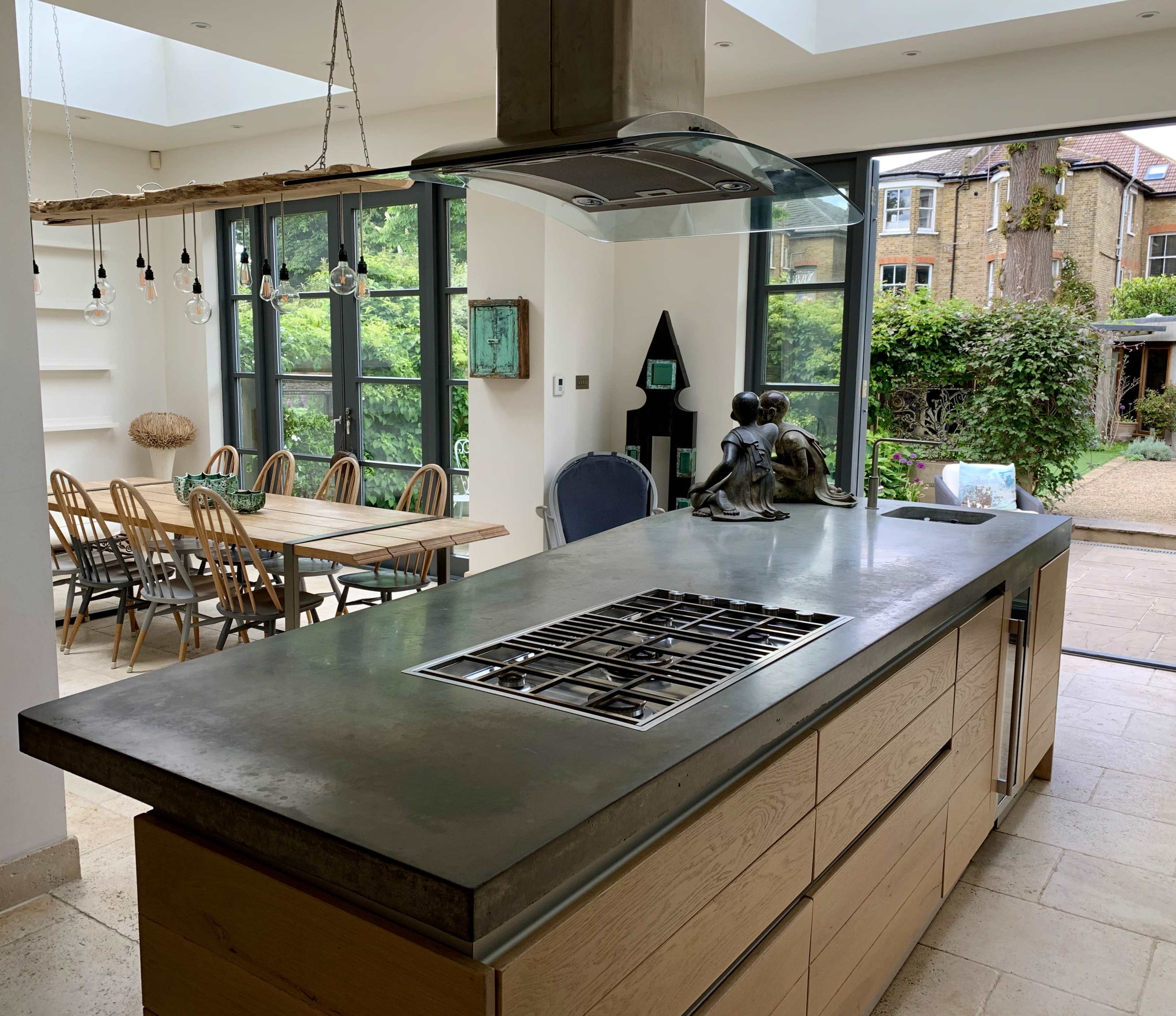 A modern kitchen features a central island with a concrete countertop and a gas cooktop, overlooking a dining area with wooden chairs and large glass doors leading to an outdoor space.