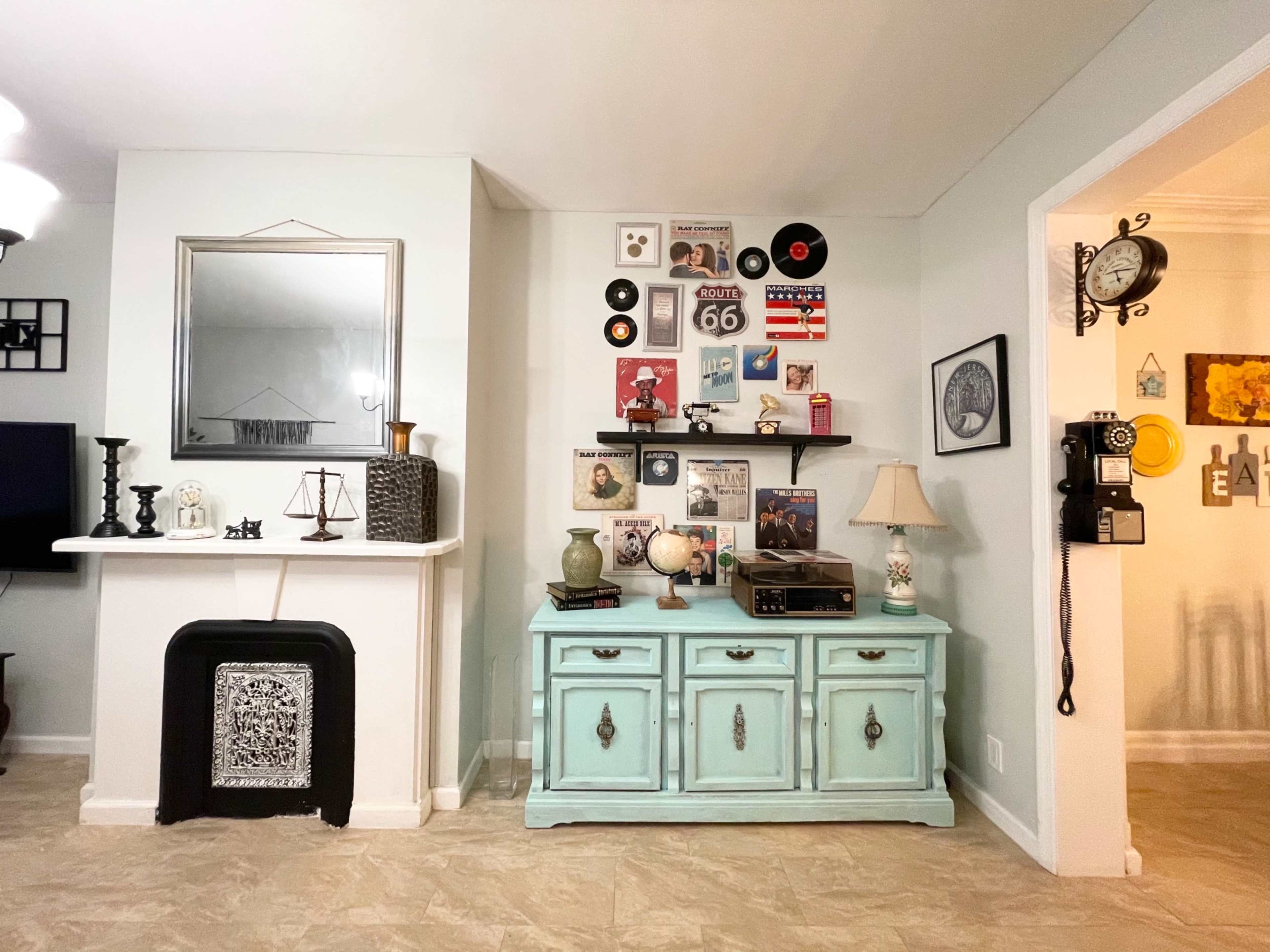 A living room with a light-colored wall featuring a vintage record and memorabilia display above a blue sideboard, while a vintage telephone hangs on the adjacent wall.