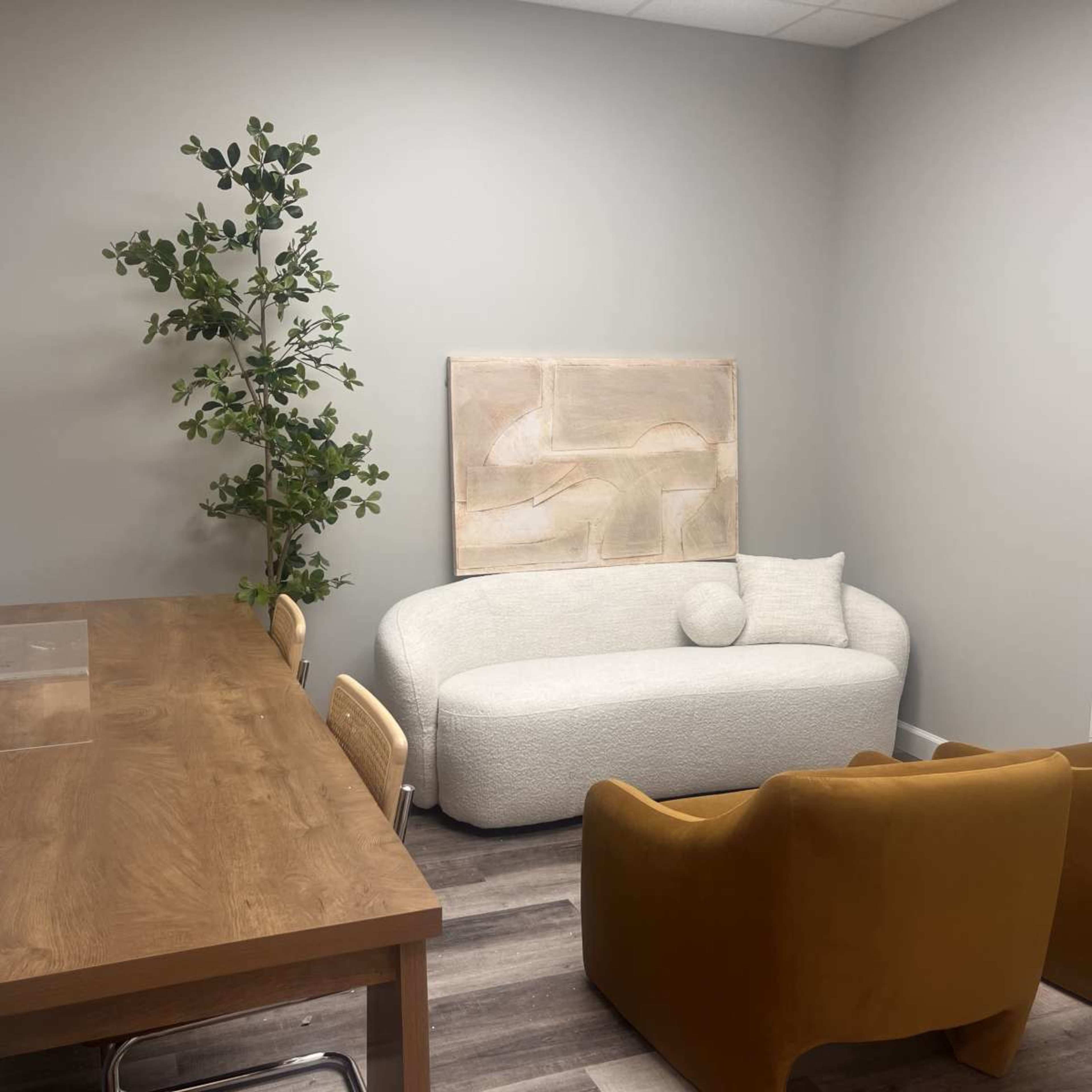 The image shows a modern interior space featuring a white curved sofa, an abstract artwork on the wall, a wooden dining table with chairs, and a tall green plant.